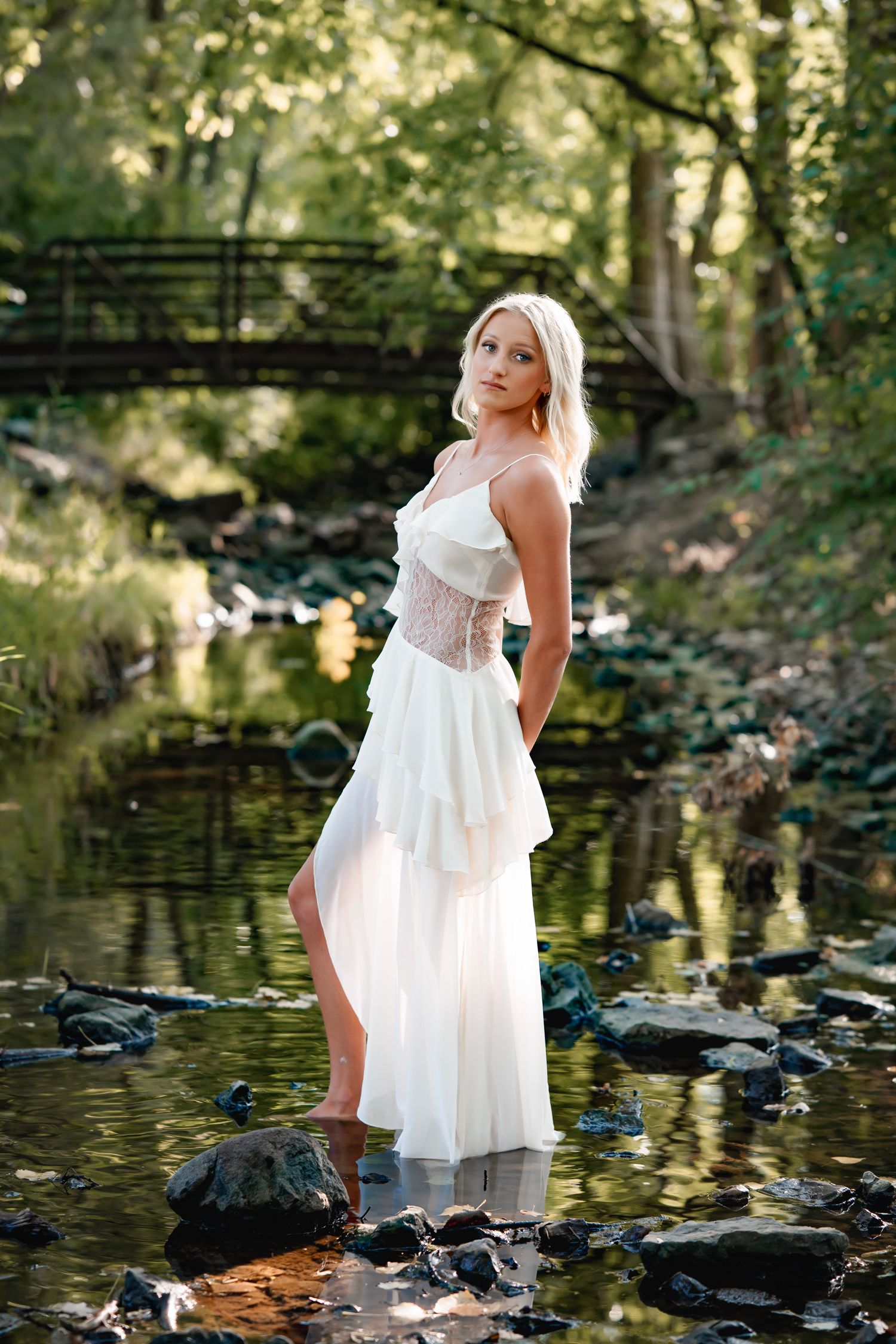 A white dress flows gracefully as someone poses by a tranquil stream with rocks and reflective water in a wooded setting.