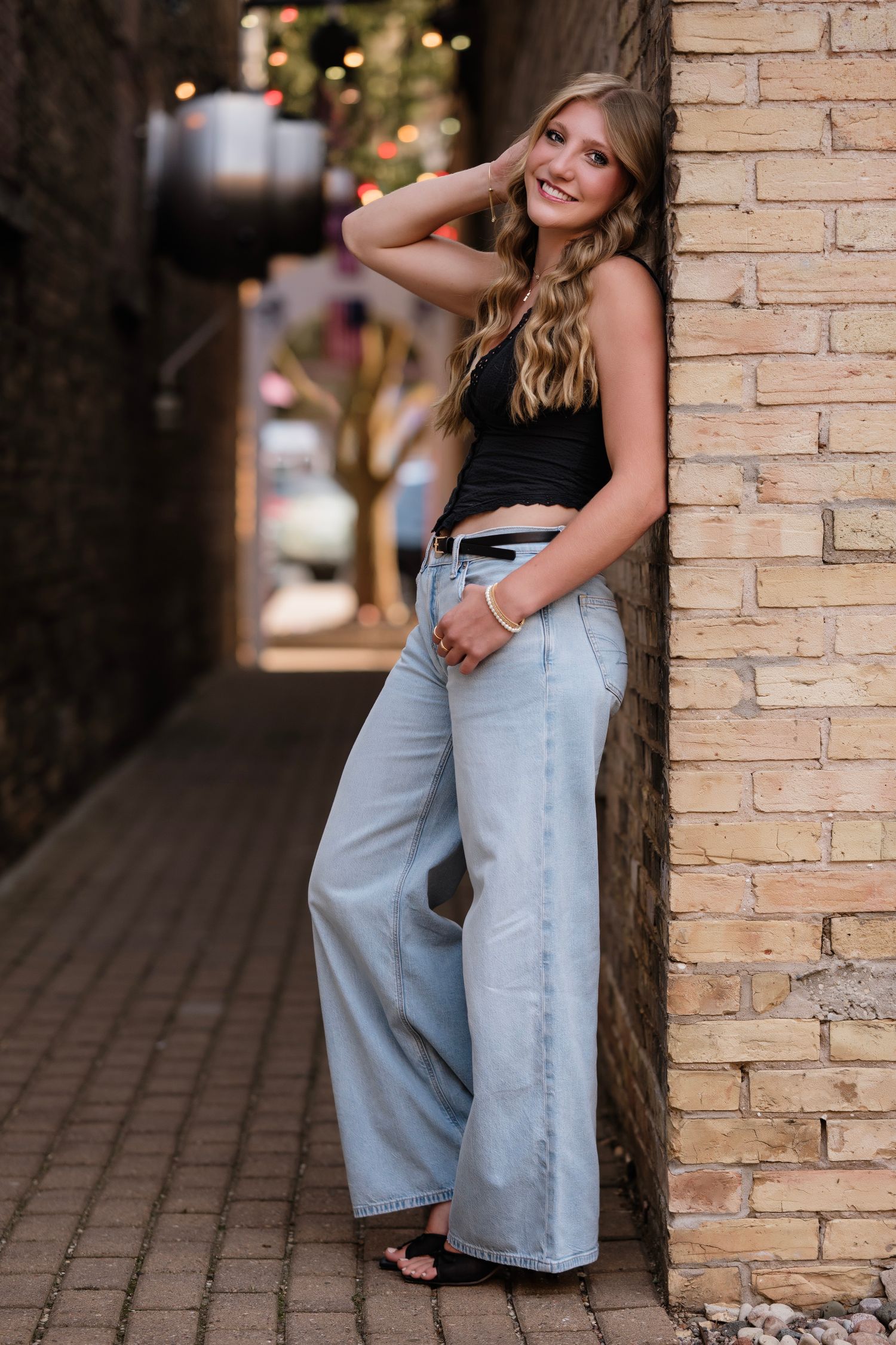Young woman in wide-leg jeans and black crop top poses against brick wall in urban alley setting.