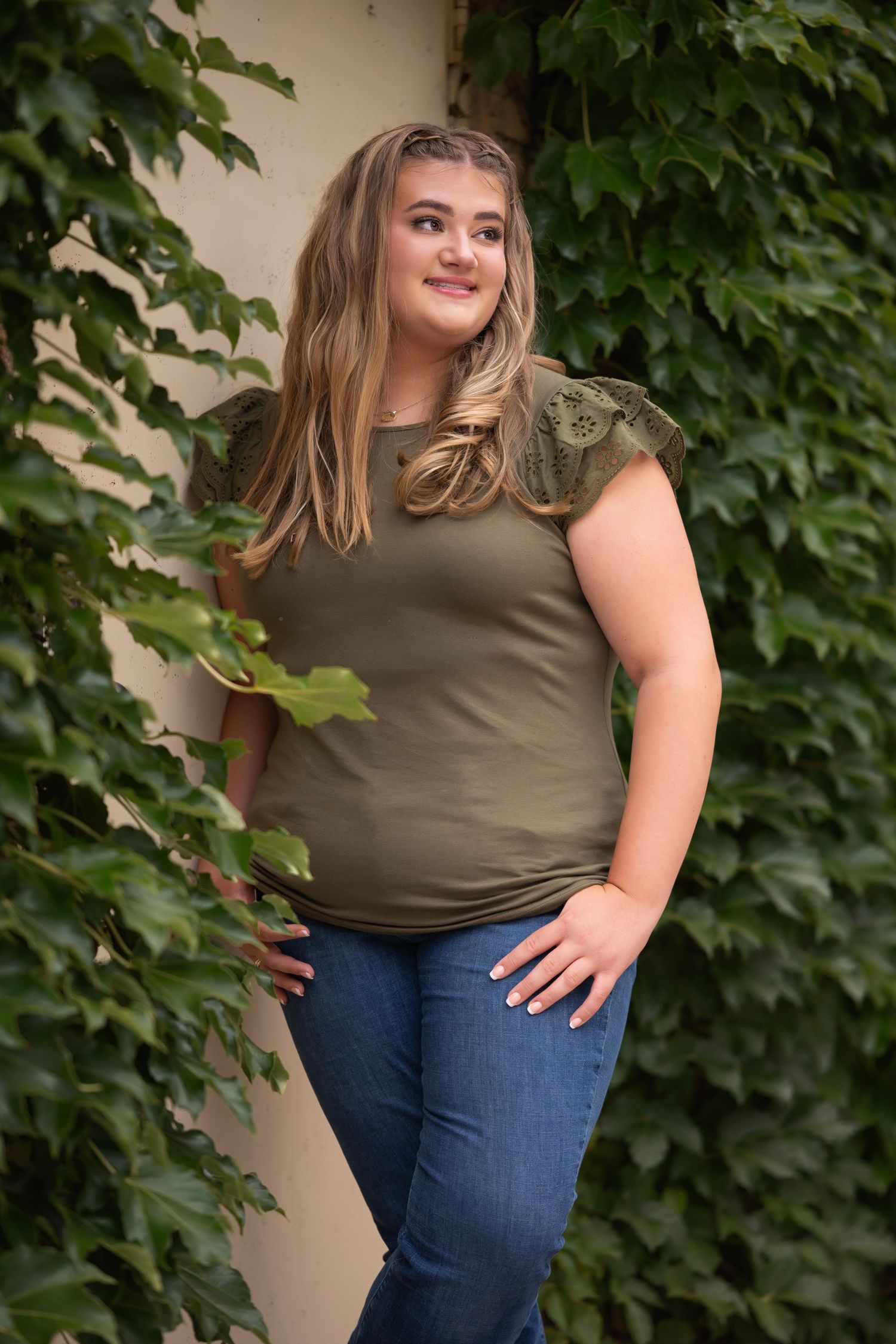 Young woman in olive green top and jeans posing outdoors against ivy-covered wall.