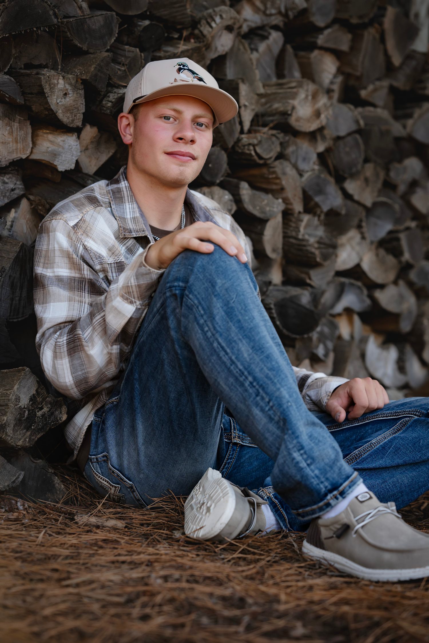 A person in plaid shirt and jeans sits against a woodpile backdrop in casual outdoor portraits.