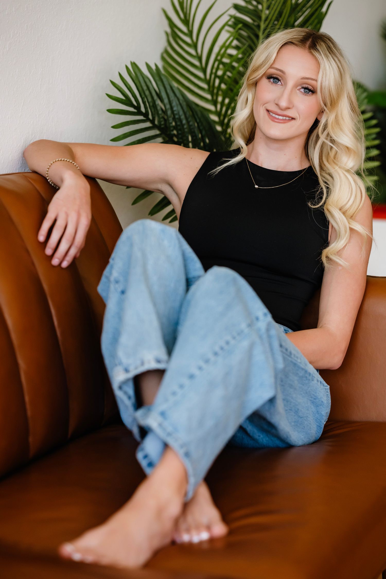 Blonde woman in black top and jeans sitting relaxed on brown leather sofa with tropical plant background.