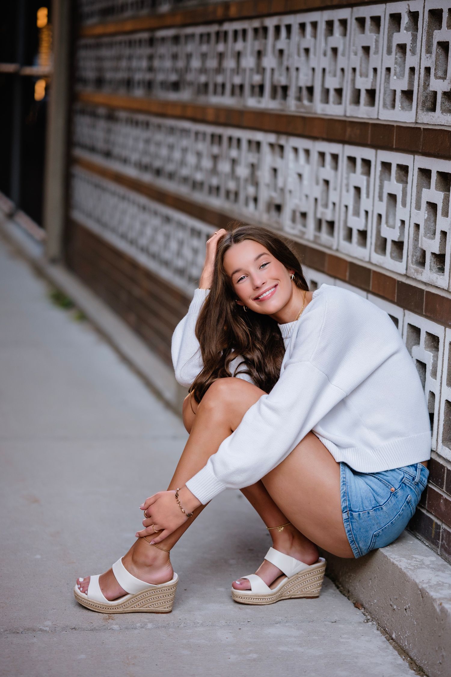 Young woman in white sweater and denim shorts sits smiling against decorative brick wall wearing wedge sandals.