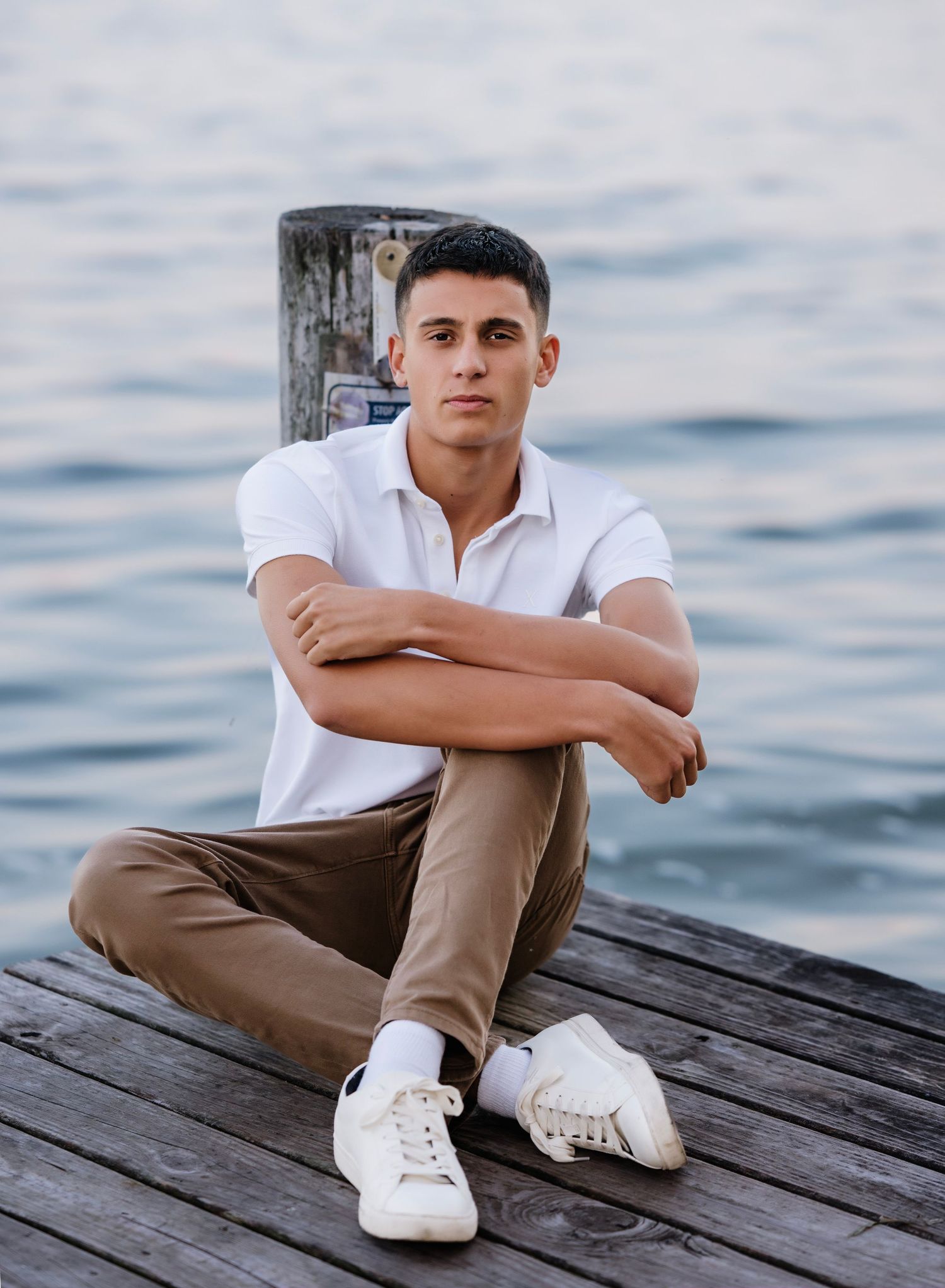 A person in a white polo shirt and khaki pants sits casually on a wooden dock by calm waters.