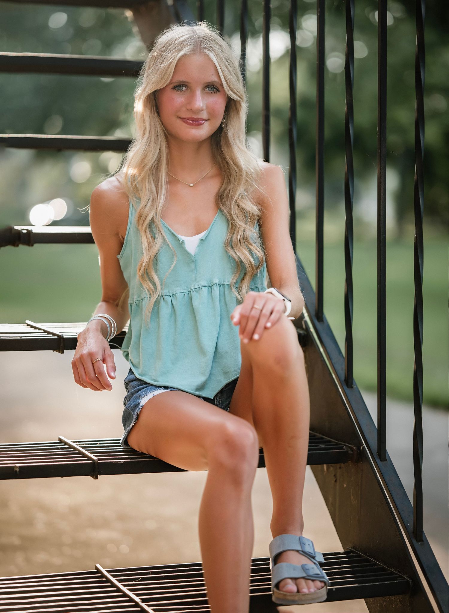 A person in a light blue sleeveless top sits on a wooden bench in an outdoor setting.