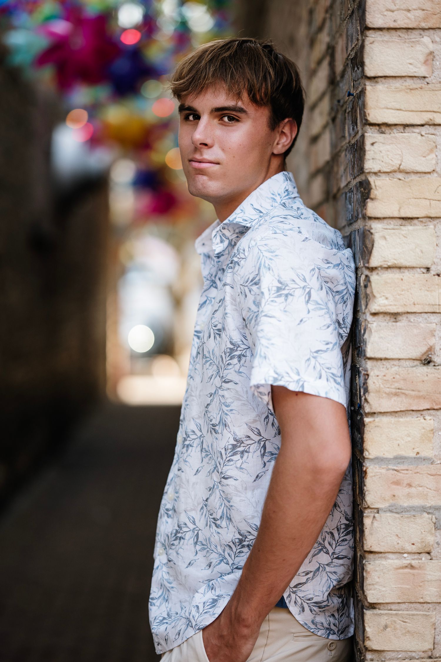 Person in white floral short-sleeve shirt poses against brick wall in outdoor setting with blurred background.