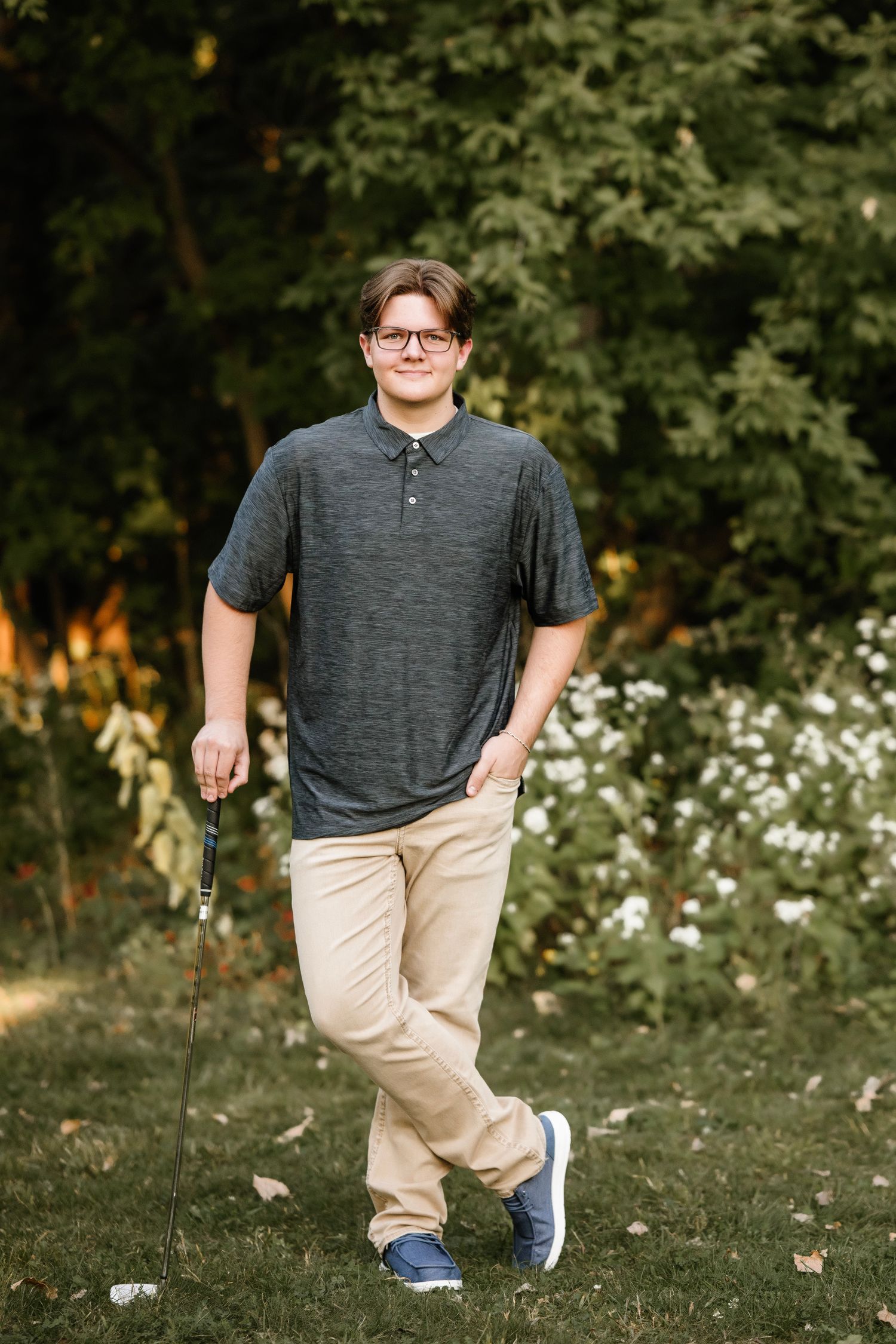 A person in a gray polo shirt and khaki pants poses outdoors in a grassy area with trees and flowers in the background.