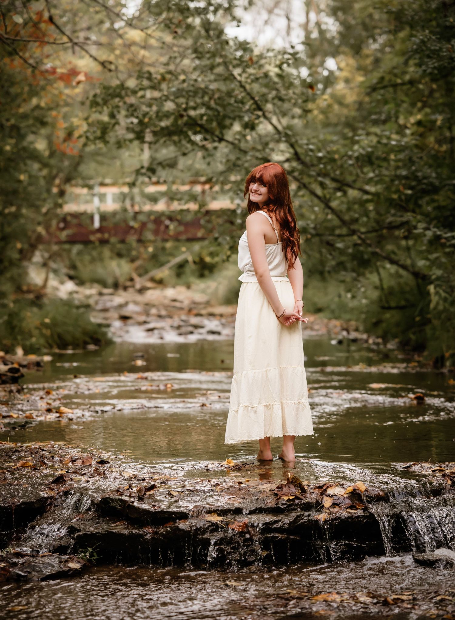 A woman in a white dress stands on rocks in a peaceful creek surrounded by lush forest scenery.