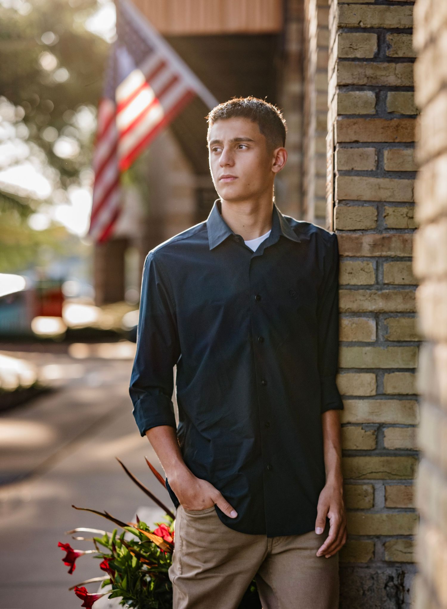A person in a navy shirt and khaki pants stands against a brick column in outdoor lighting with an American flag visible.