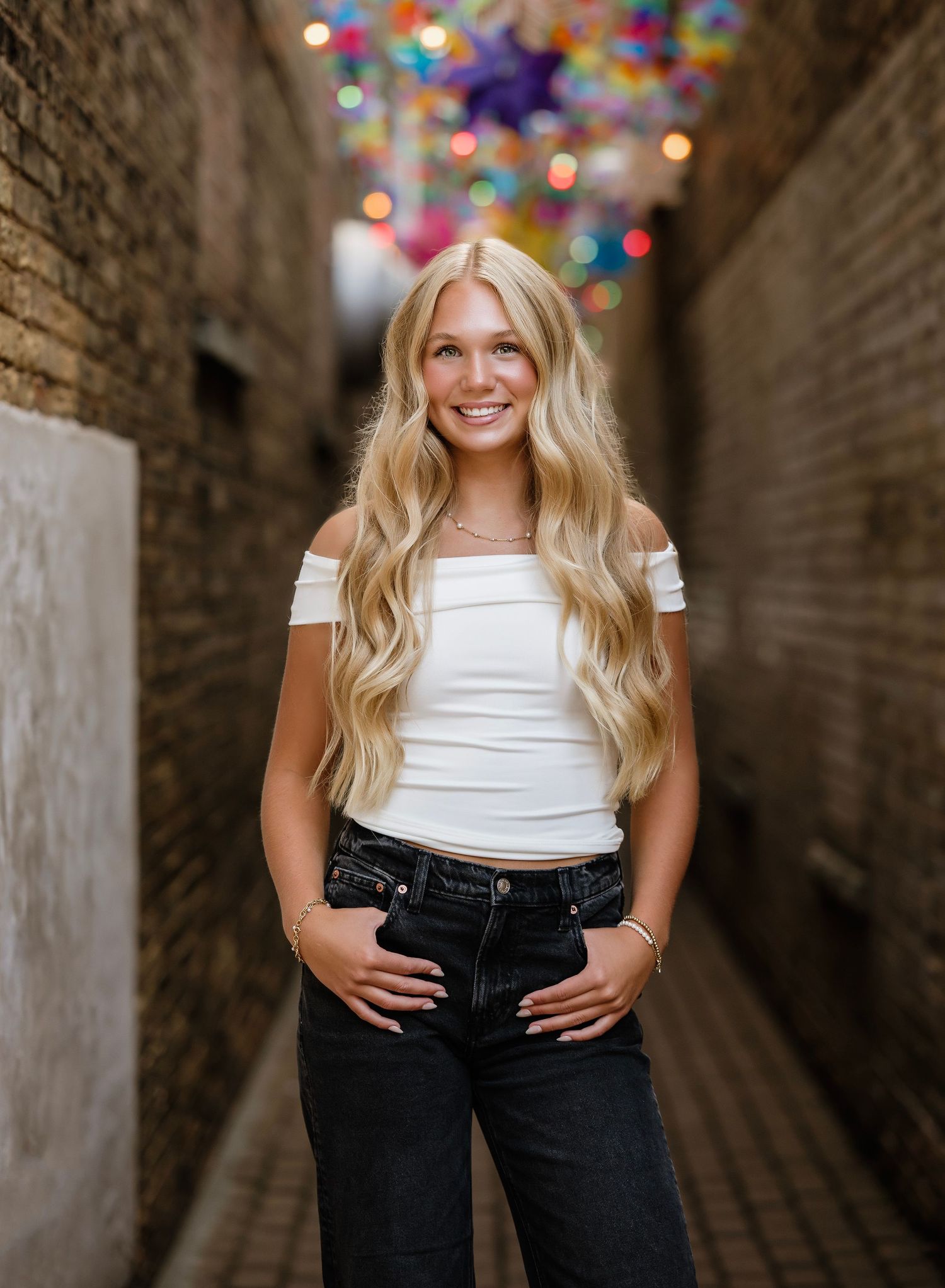 A person in a white top and dark jeans poses in a brick tunnel with colorful bokeh lights in the background.