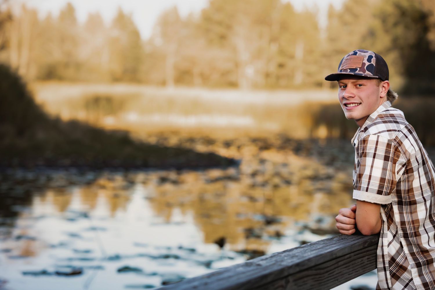 A person in a plaid shirt sits on a wooden dock by a serene lake during golden hour.