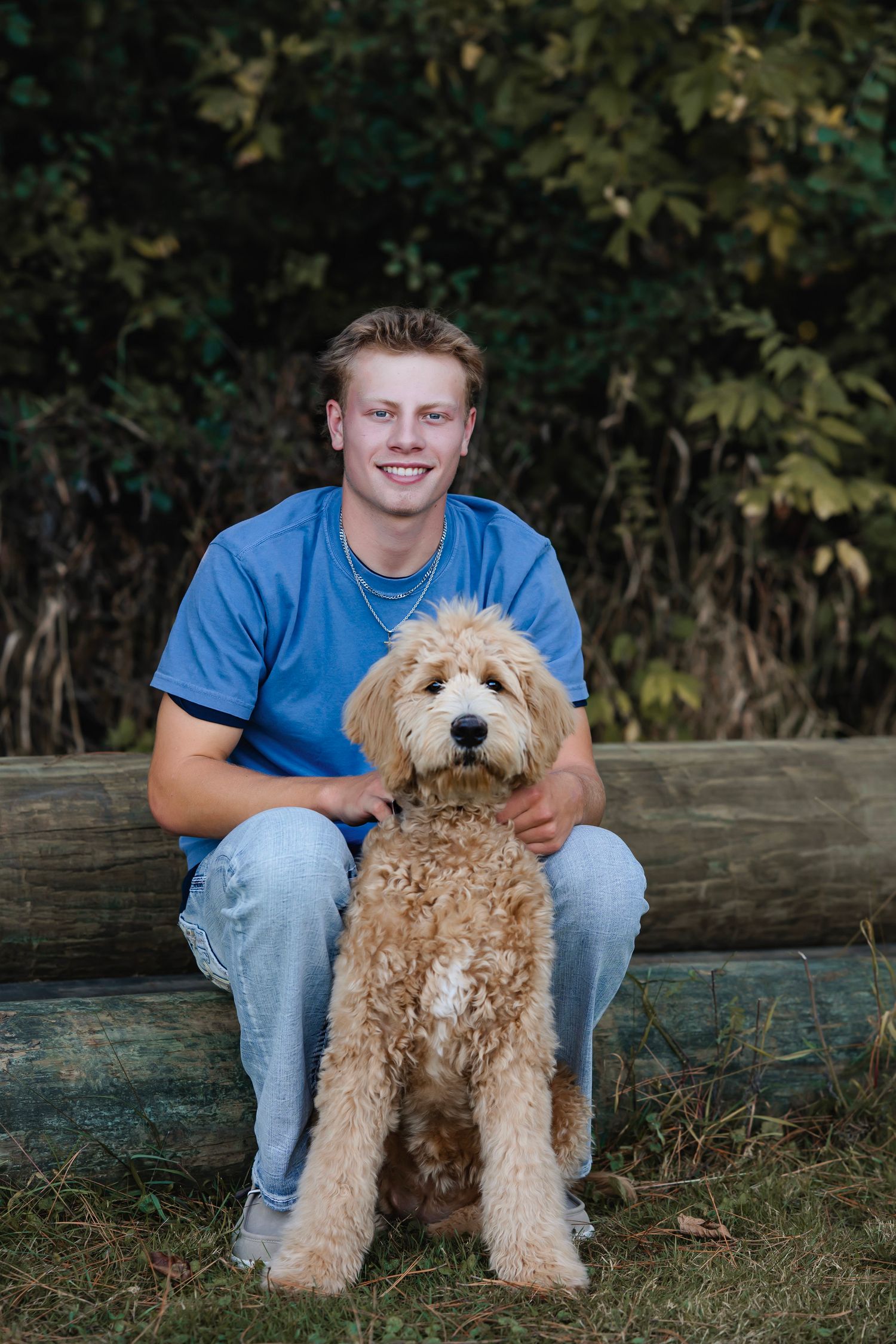 A fluffy goldendoodle puppy sits on a wooden bench with someone in blue jeans and a t-shirt in an outdoor setting.