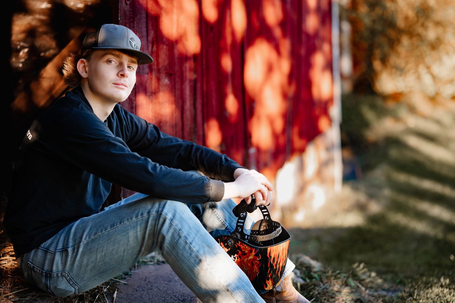 Someone in a baseball cap and denim sits against a red wall outdoors with autumn lighting and shadows.