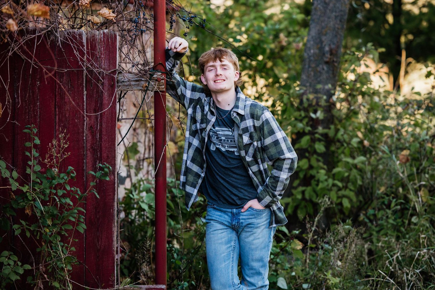 A person in a plaid shirt and jeans poses casually against a wooden post in an outdoor garden setting.