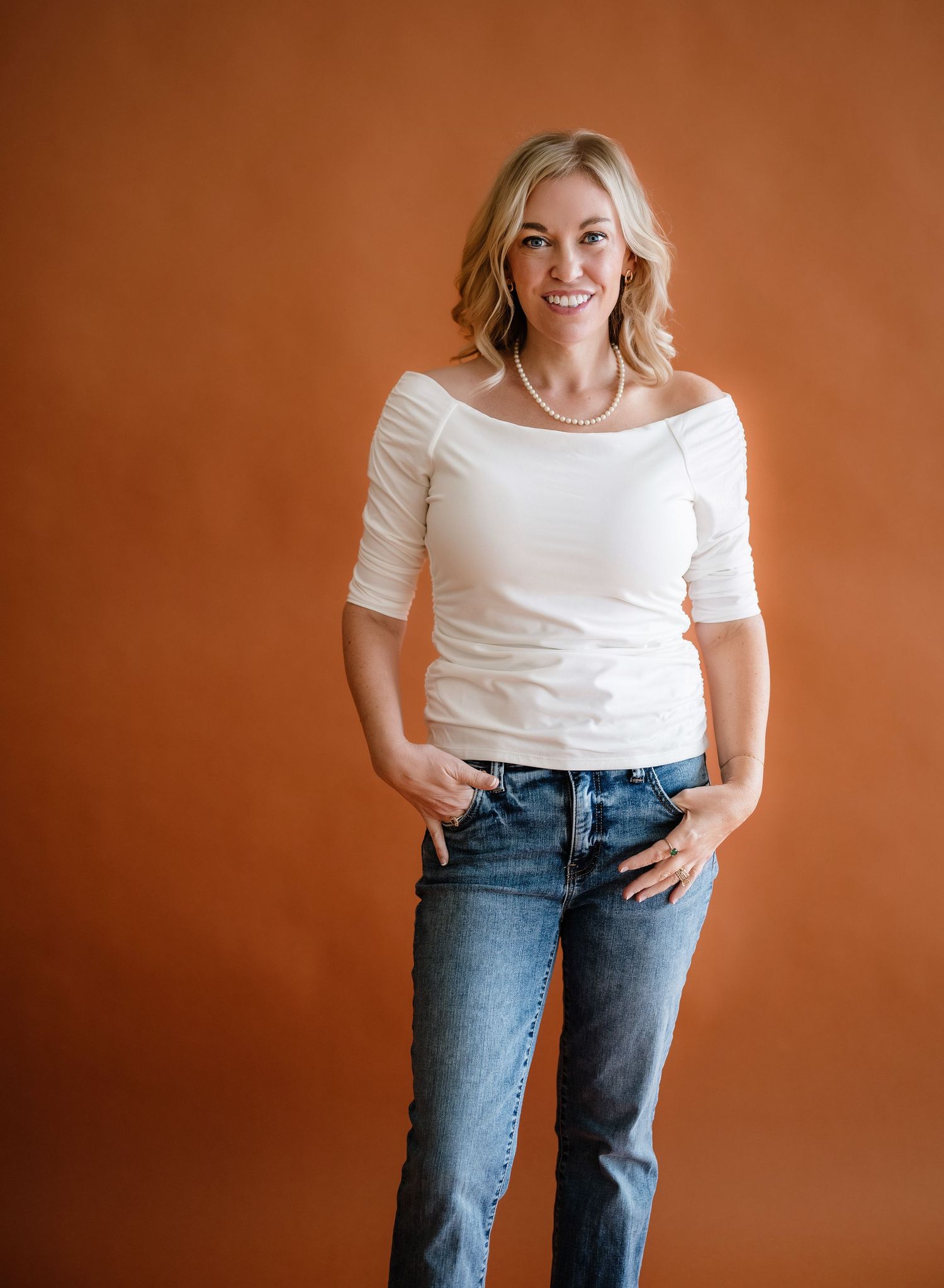 Casual portrait against brown background showing person in white off-shoulder top and blue jeans with relaxed pose.
