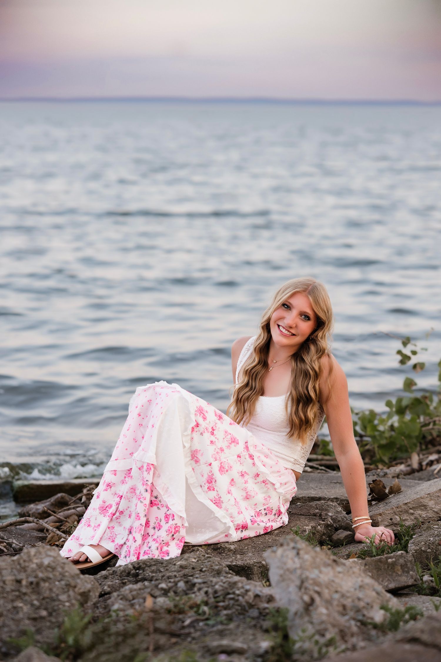 Young woman with long blonde hair sits on rocks by the ocean in a white and pink floral maxi dress, smiling.