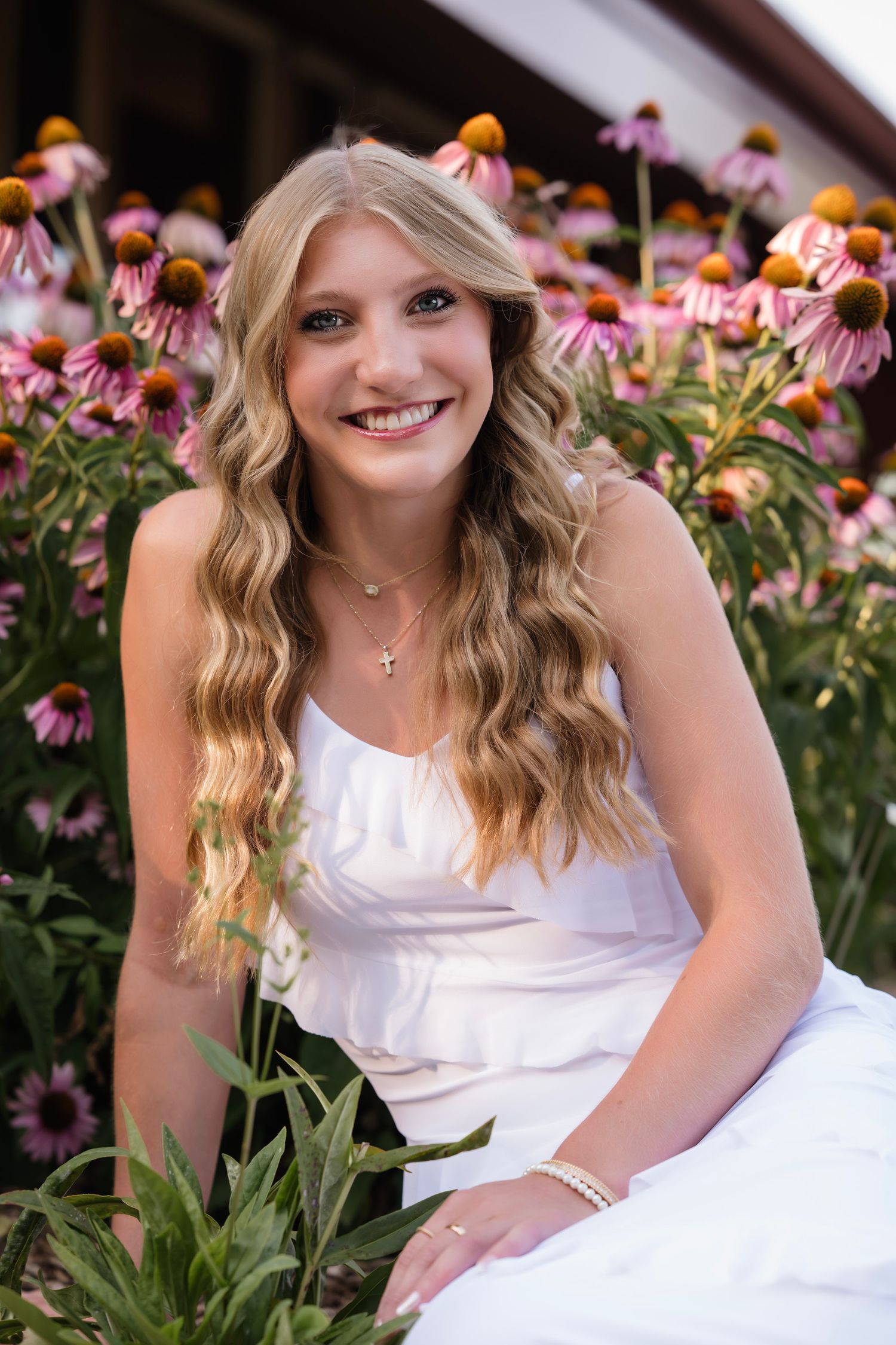 Smiling young woman in white dress sitting among pink coneflowers, wearing a cross necklace and bracelet.