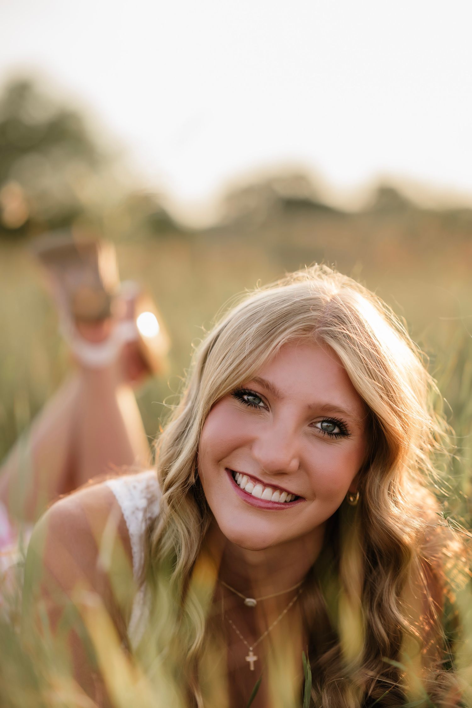 Young blonde woman smiling in a golden field at sunset, wearing a cross necklace, lying in tall grass.