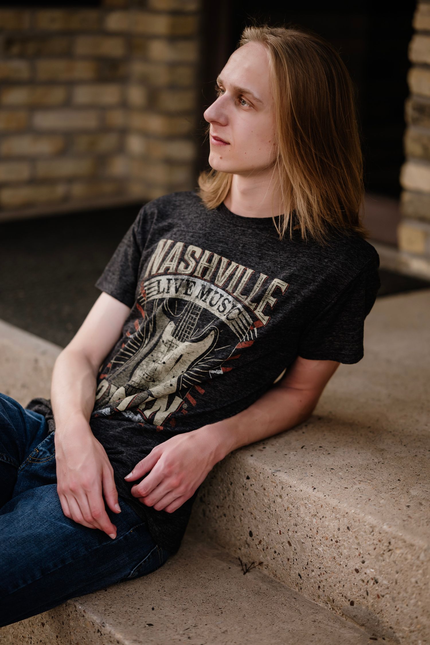 Young man with long blonde hair sitting on stone steps wearing a Nashville Live Music graphic tee and jeans.