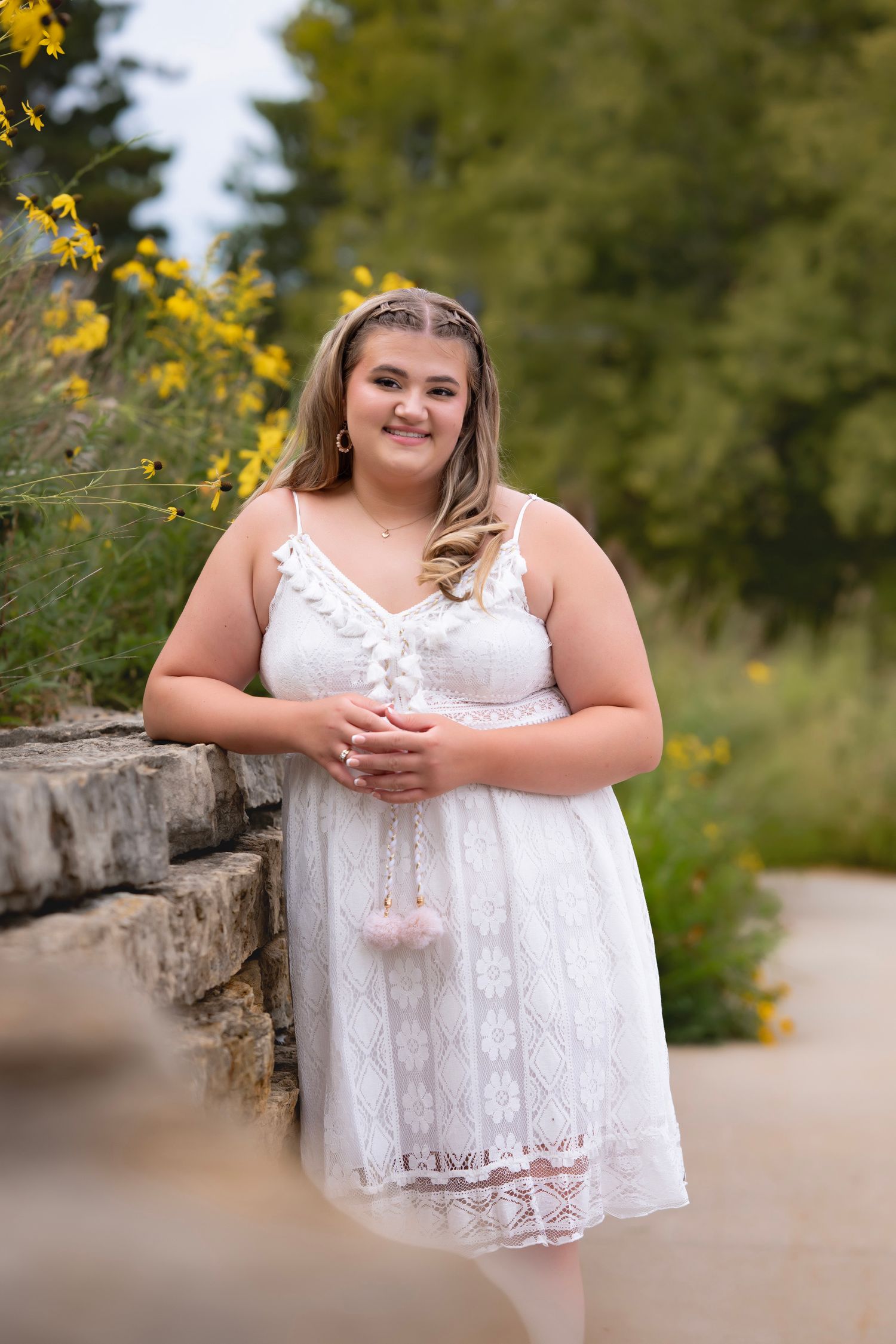 Young woman in white lace dress posing outdoors near stone wall with yellow wildflowers and green trees.