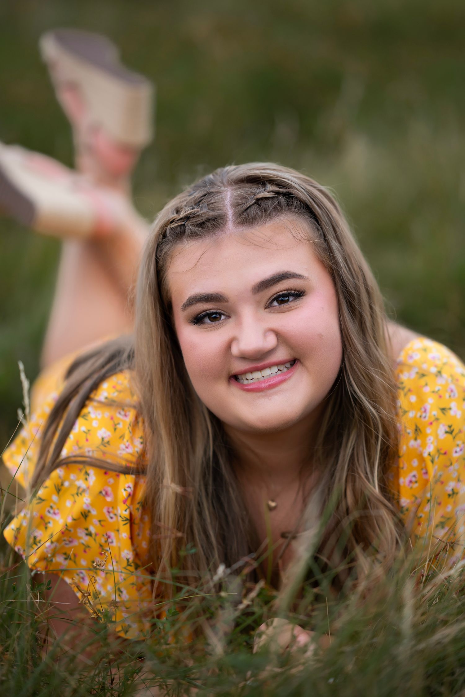 Smiling teenage girl with braces lying in grass wearing a yellow floral dress for senior portrait photos.