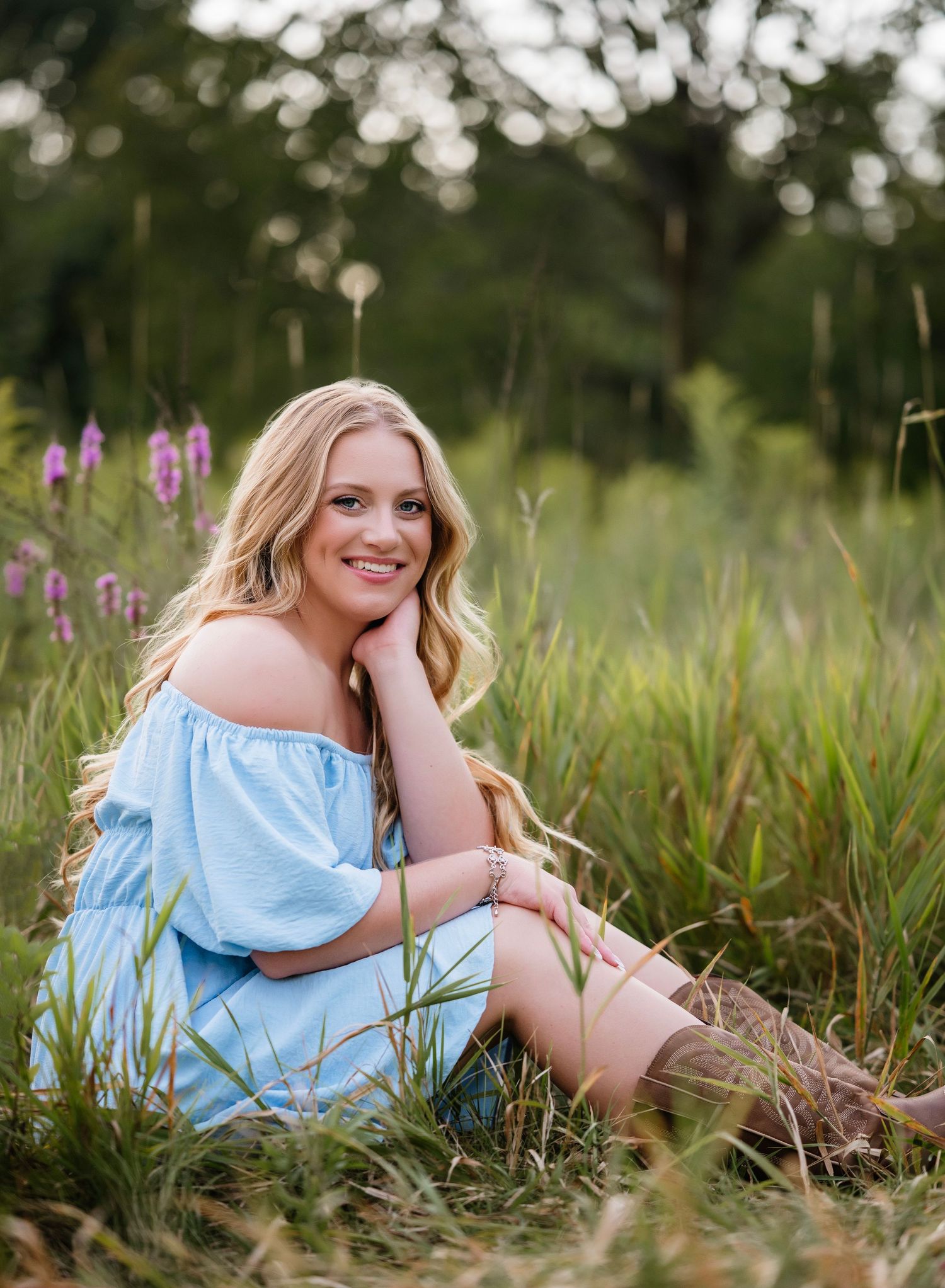 A person in a light blue dress sits in a grassy meadow with purple wildflowers on a summer evening.