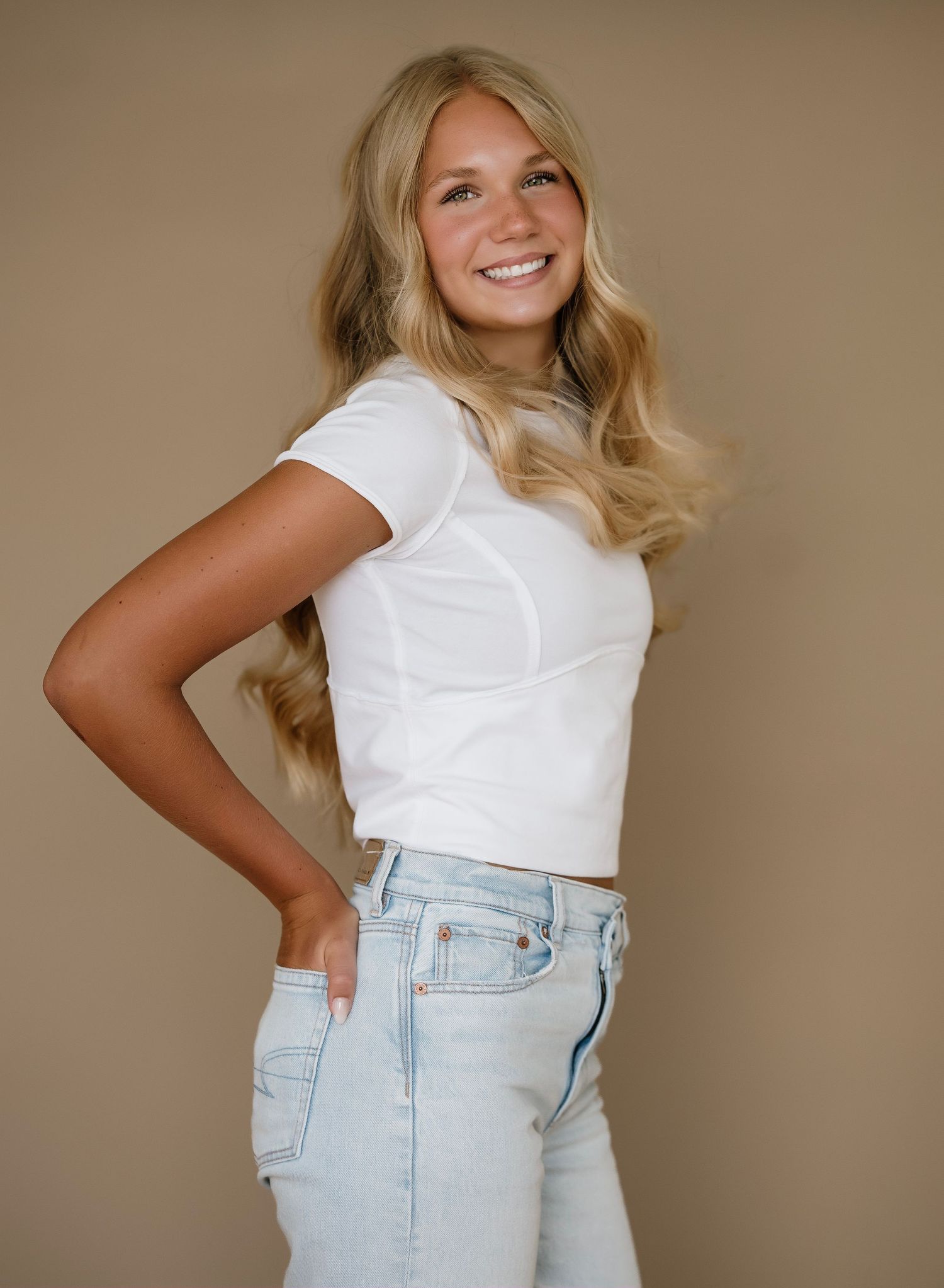 Portrait of someone in a white crop top t-shirt and light wash jeans posing against a neutral beige background.