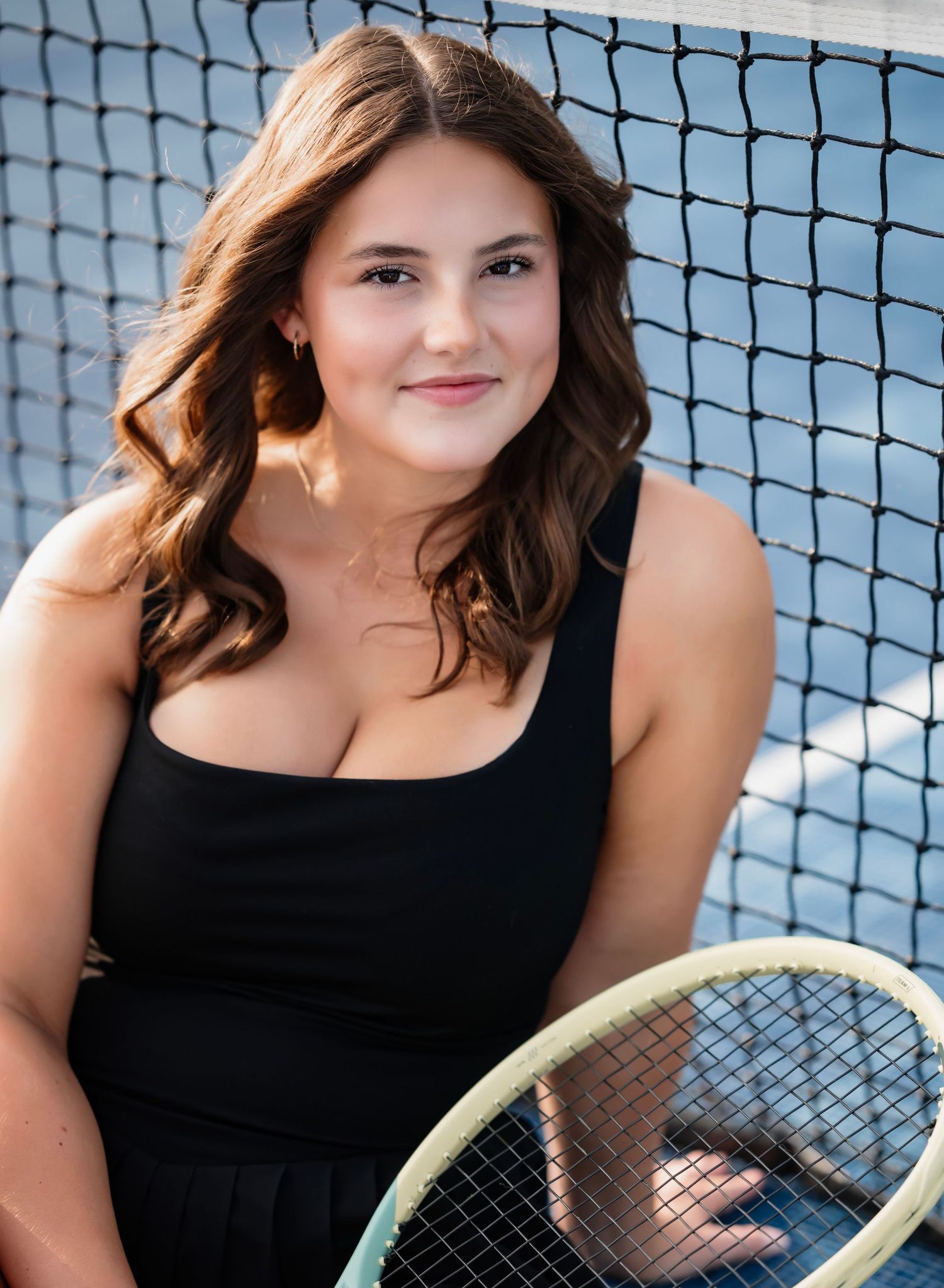 Tennis player poses with racquet against net background in black tank top during outdoor portrait session.