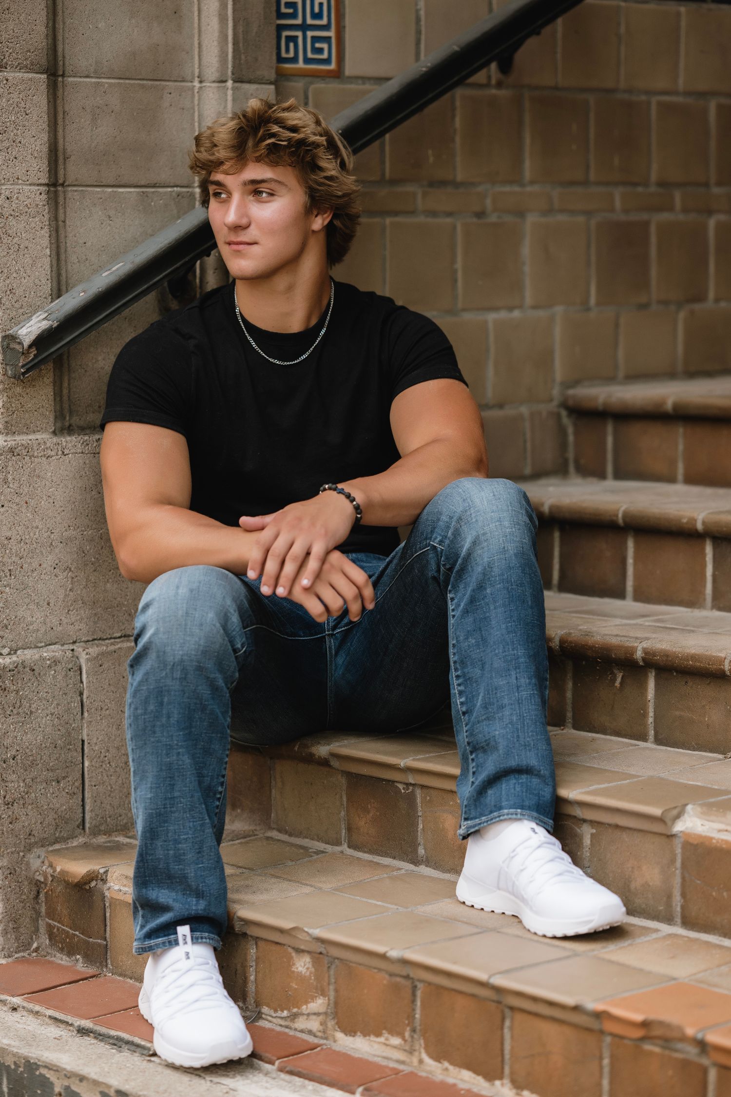A person in black shirt and blue jeans sits casually on concrete steps wearing white sneakers.