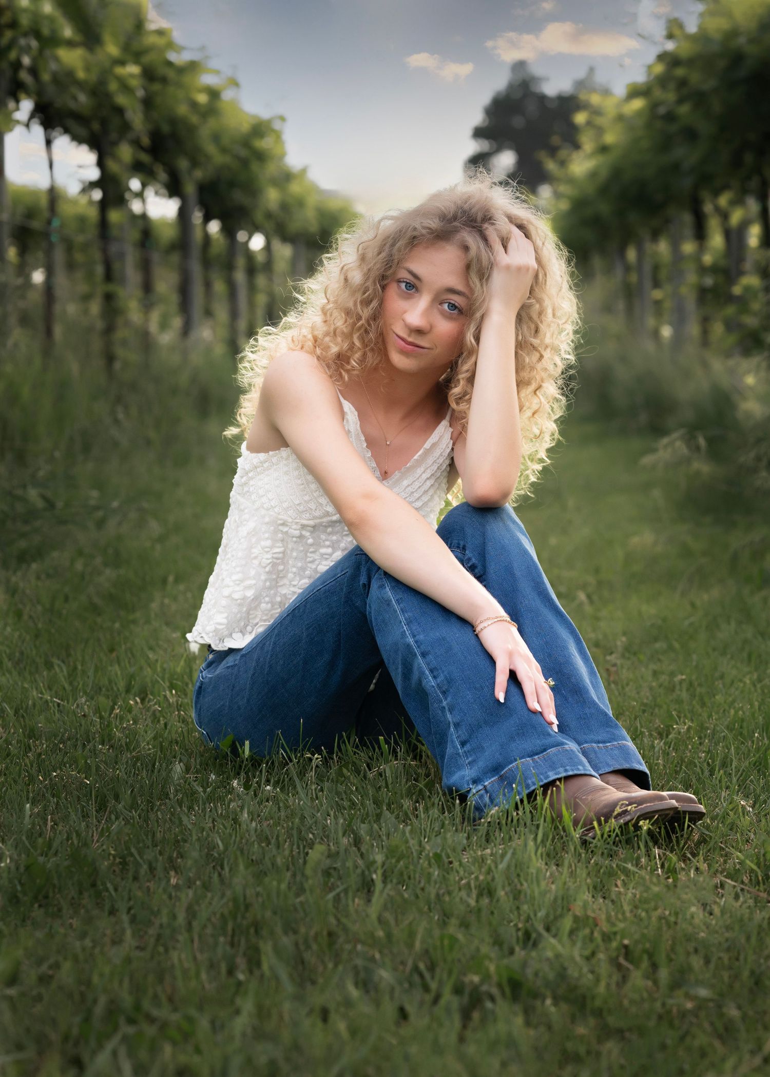 Young woman with curly blonde hair sits on grass in vineyard wearing white top and jeans.