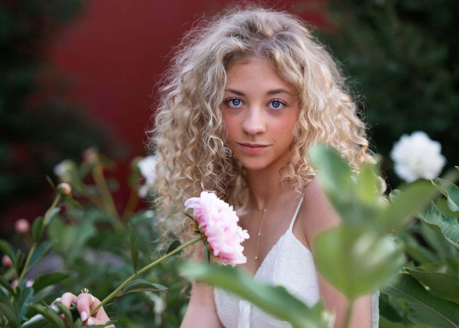 Young girl with curly blonde hair poses among pink and white blooming flowers in a garden.