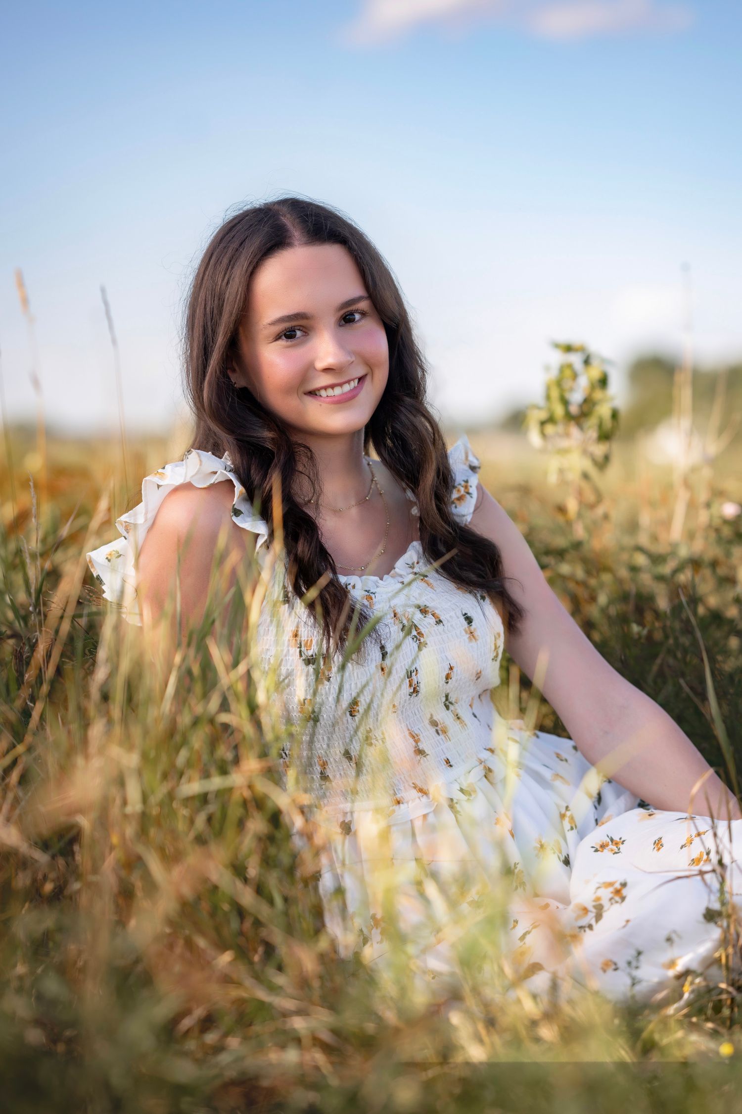 Smiling young girl in a white floral dress sitting in a golden meadow field on a sunny day.