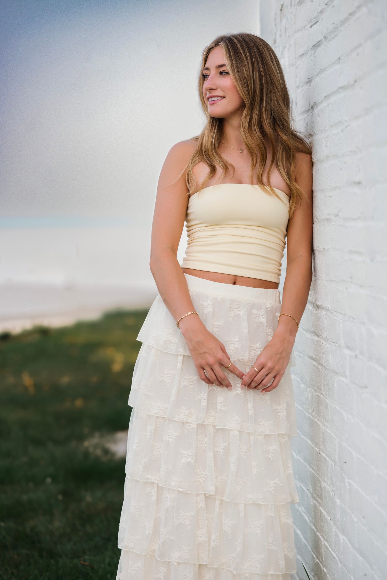 Young woman in cream tube top and tiered ruffle skirt leaning against white brick wall outdoors.