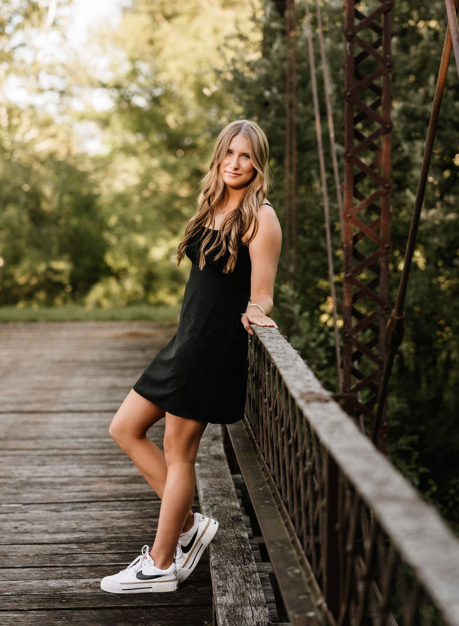 Young blonde woman in black mini dress and Nike sneakers posing on a wooden bridge surrounded by lush greenery.