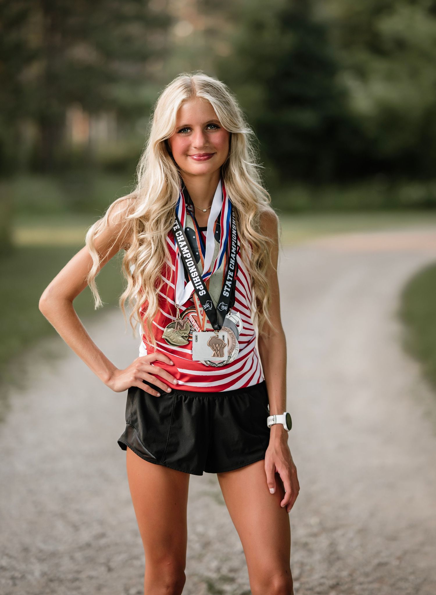 An athlete wearing patriotic red white and blue attire poses with medals on a gravel path outdoors.