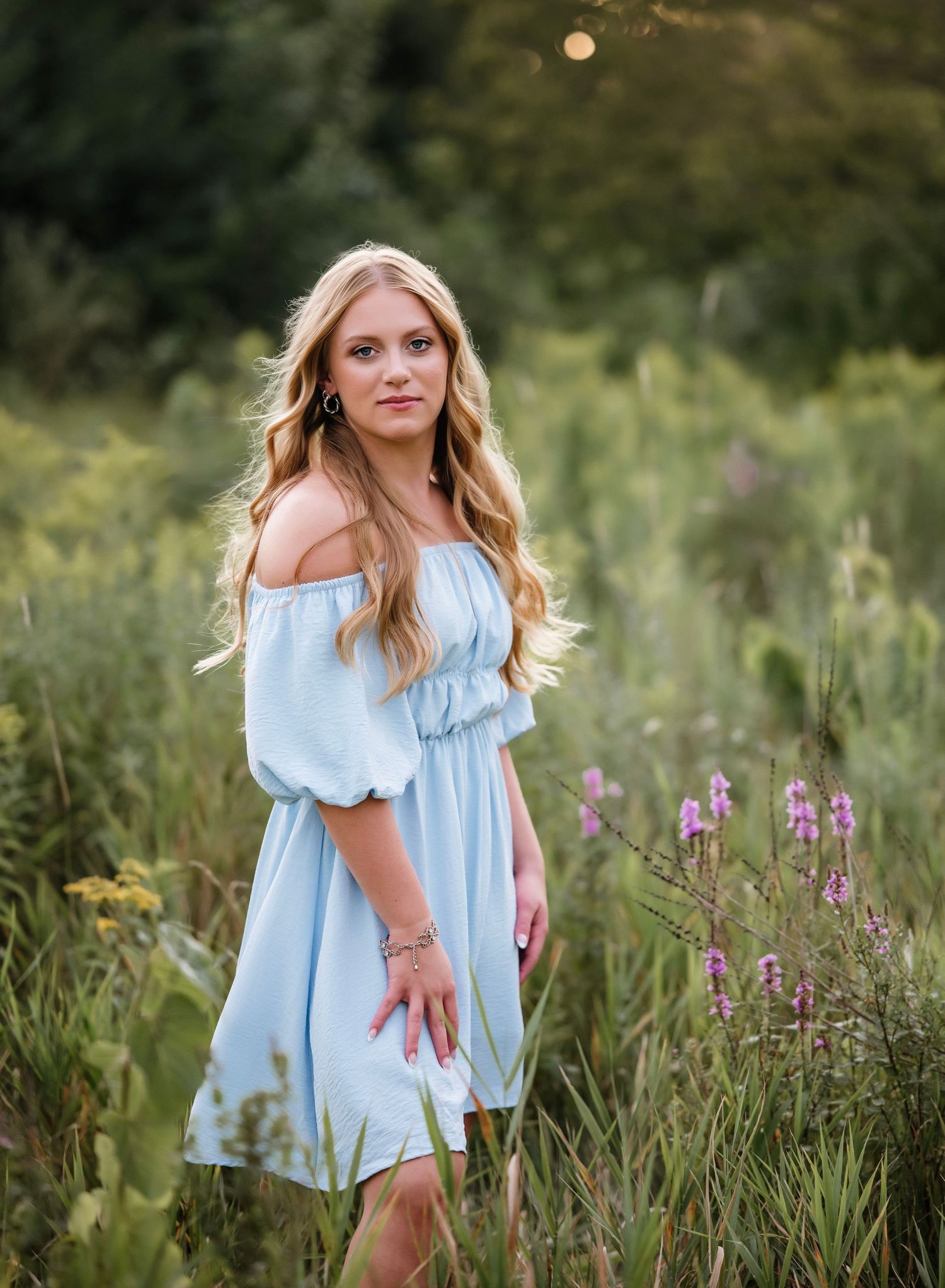 Young blonde woman in light blue off-shoulder dress poses gracefully in a wildflower meadow during golden hour.