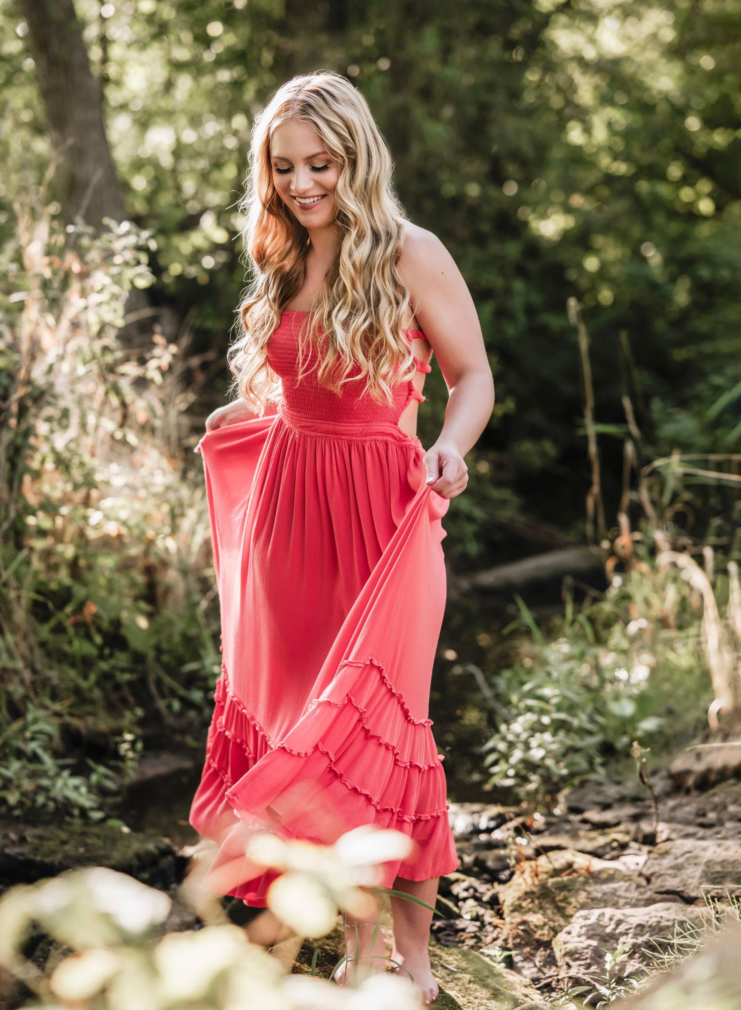 A person in a flowing coral dress poses gracefully on a forest path surrounded by lush greenery and dappled sunlight.