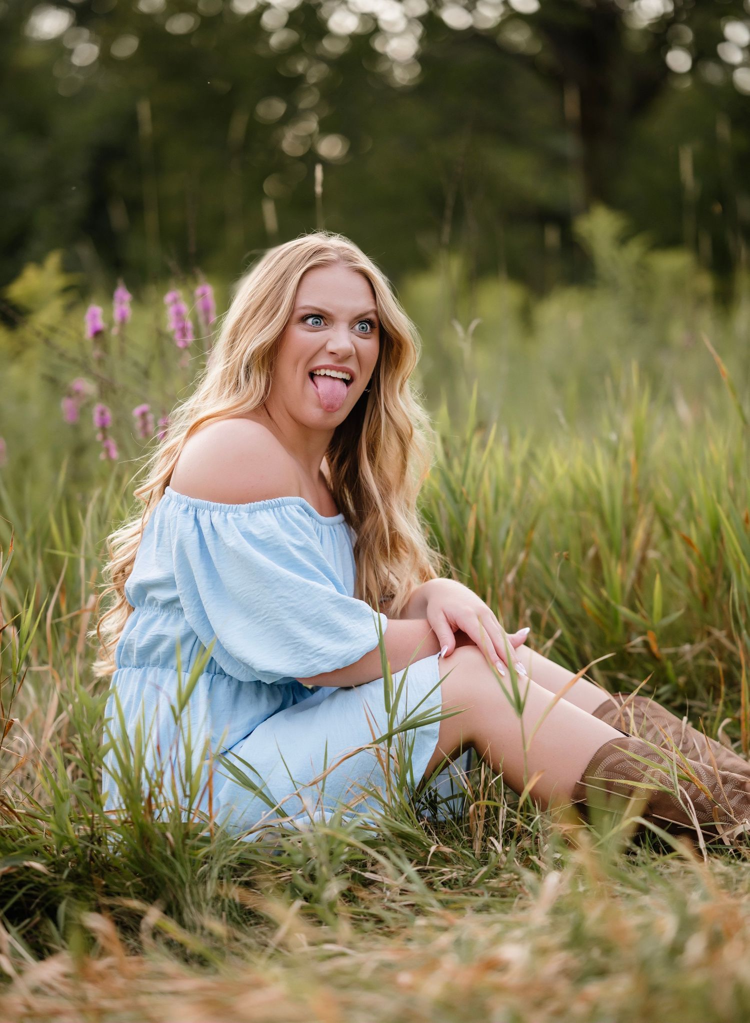 A person in a light blue top lies in a grassy meadow with purple wildflowers on a sunny summer day.