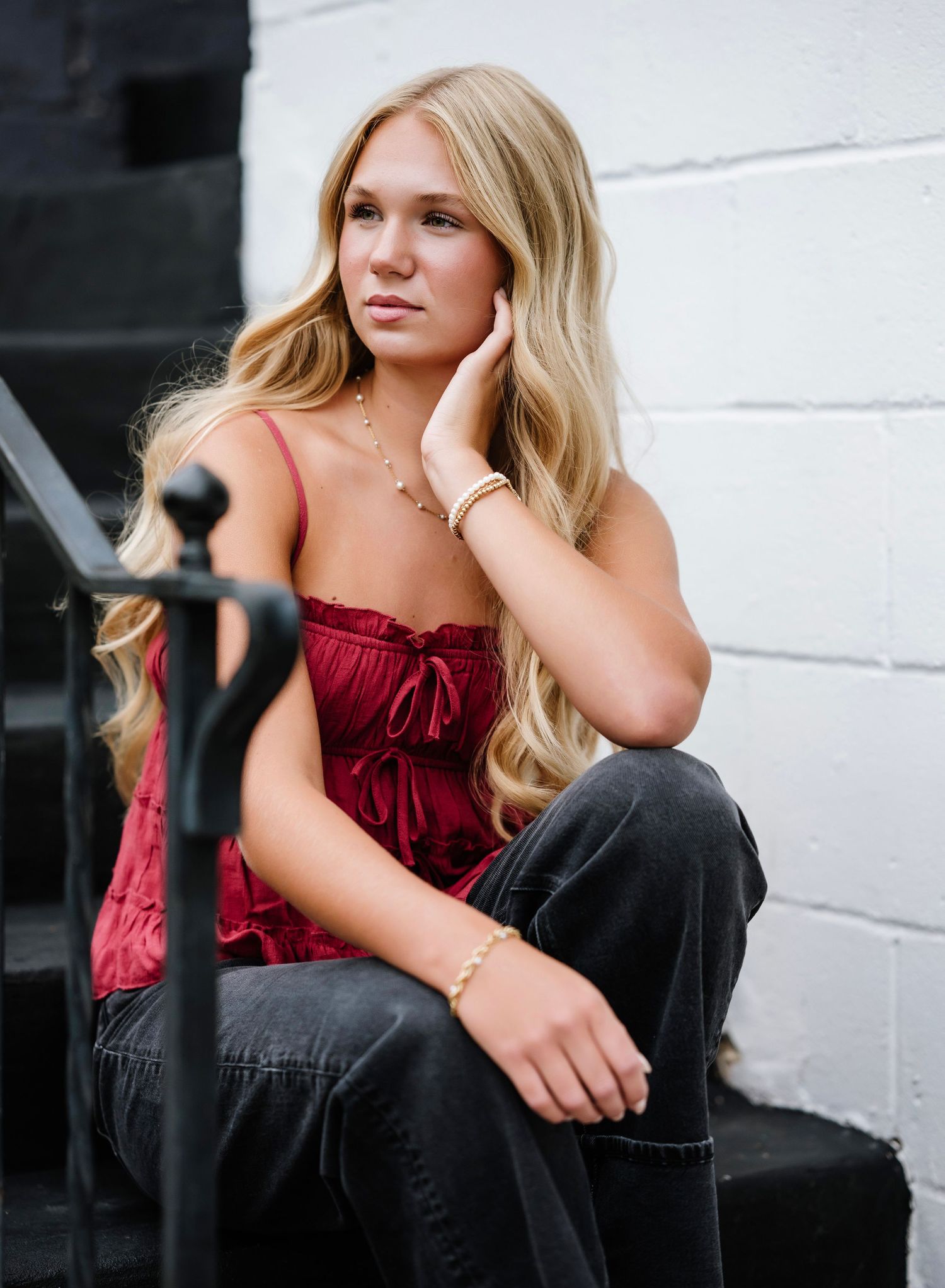 A person in a red velvet top and dark pants sits casually on black steps against a white wall.