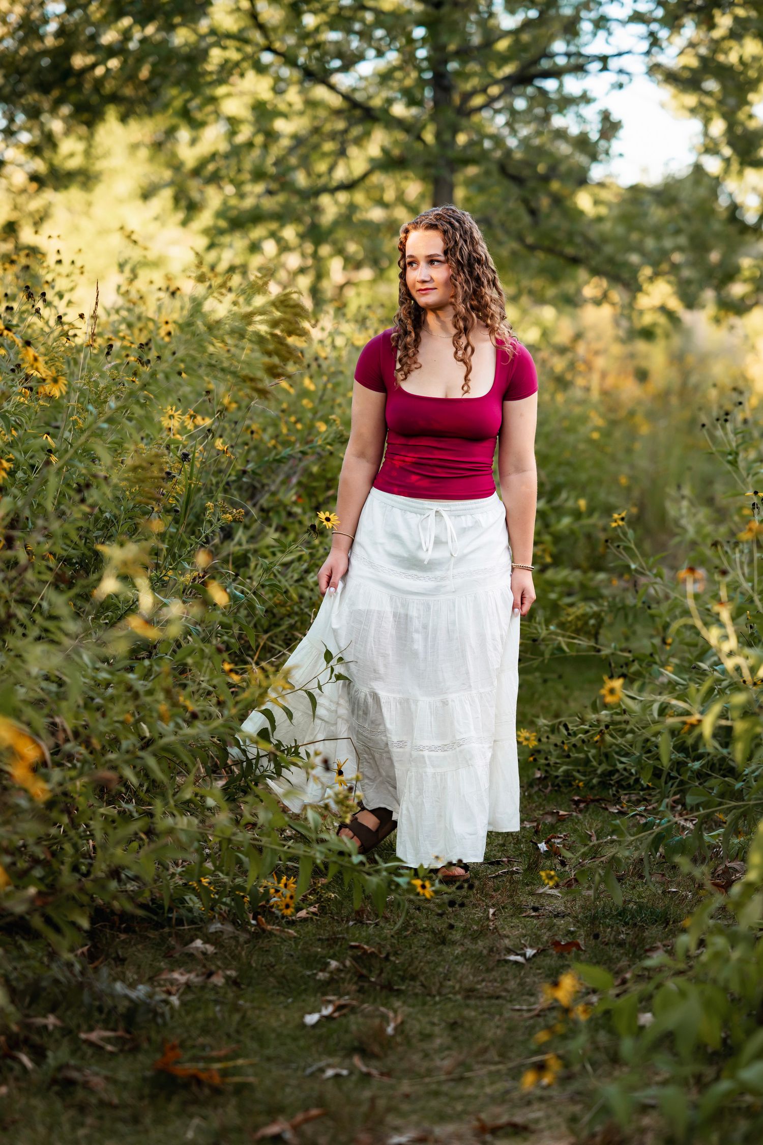 A figure in a red top and white skirt stands among flowering bushes in a sunlit garden setting.