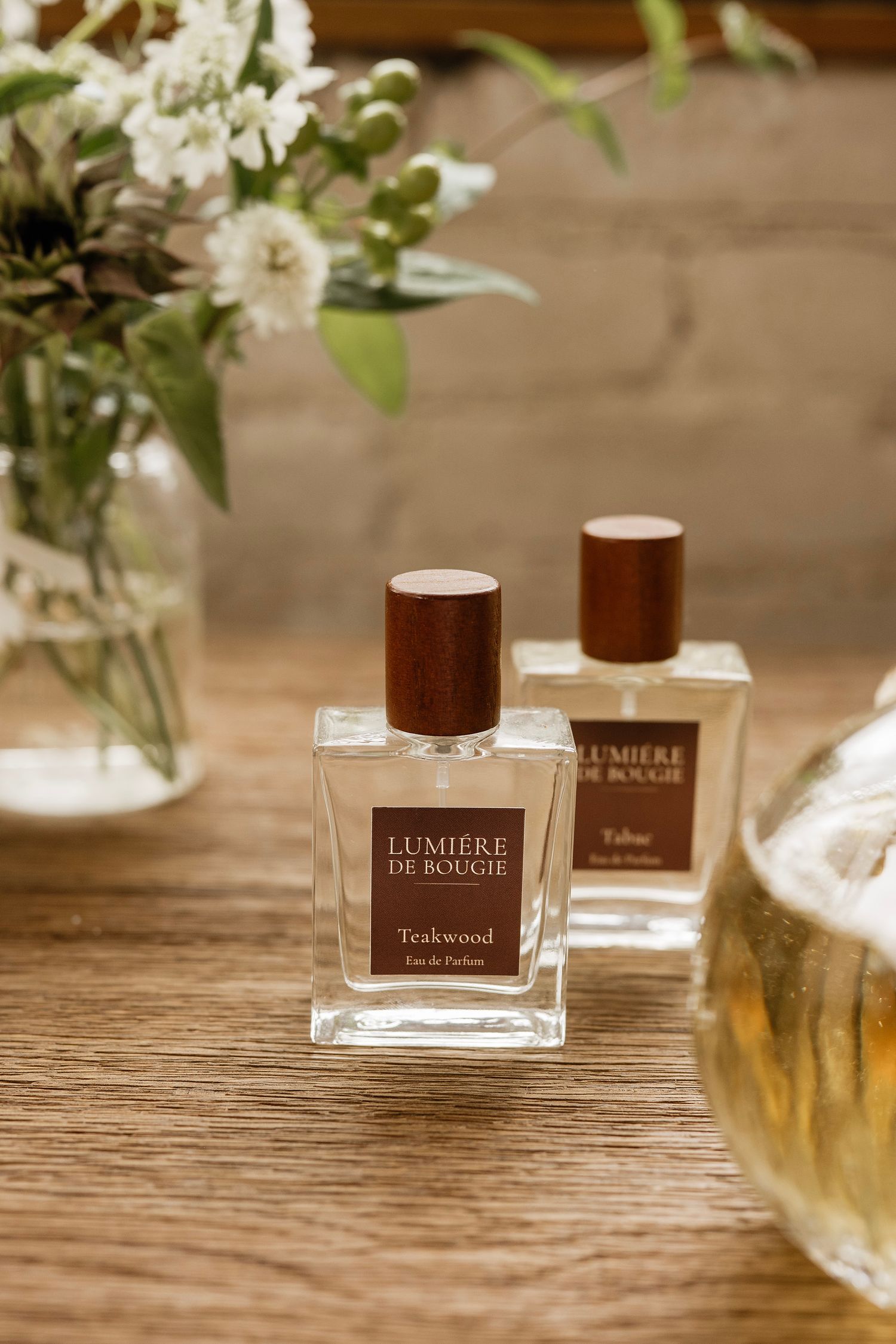 Two Lumière de Bougie perfume bottles, Teakwood and Tobacco, displayed on a wooden table with white flowers in the background.