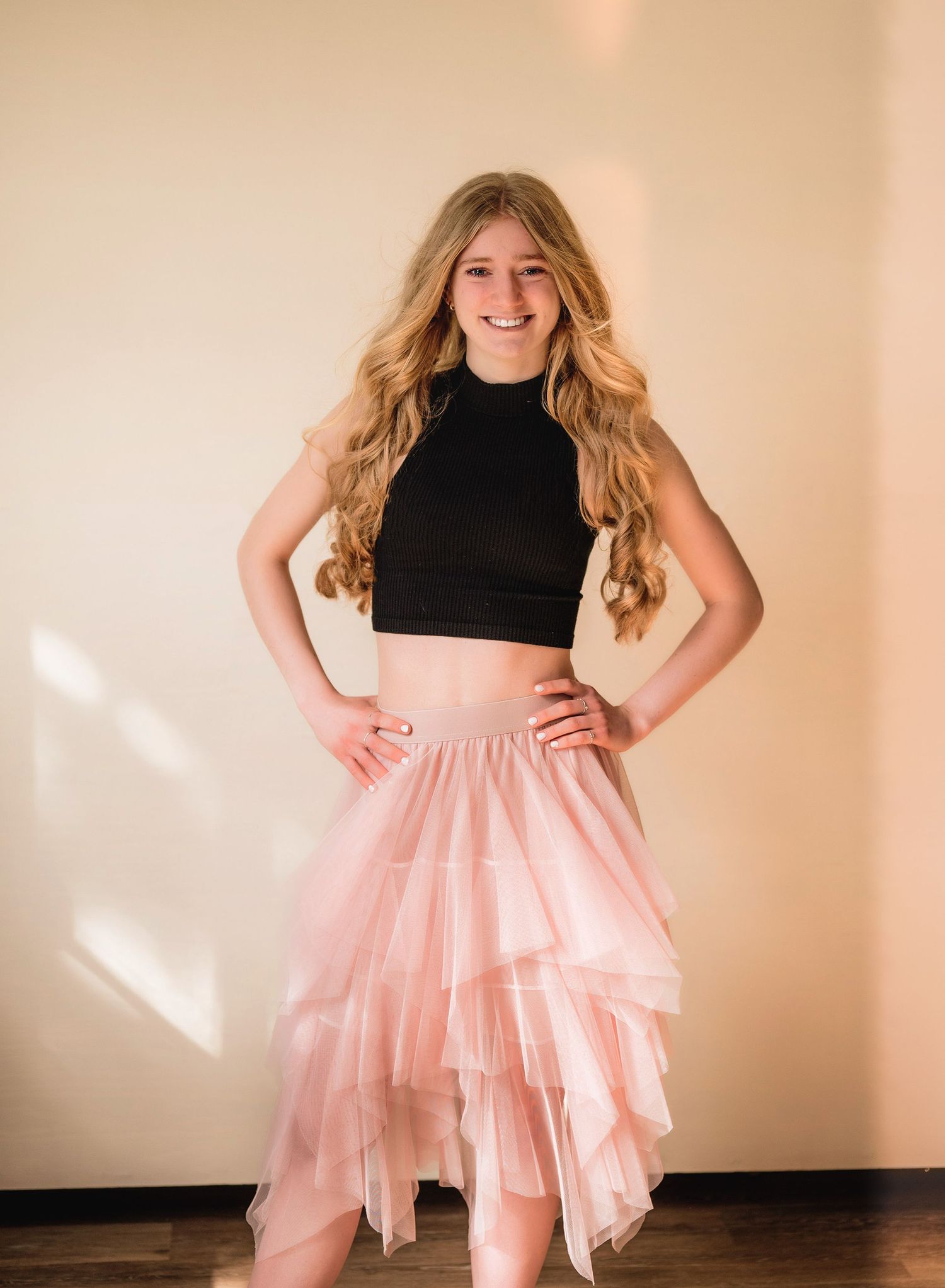 Smiling teenage girl in black crop top and pink tulle skirt posing with hands on hips against beige wall.
