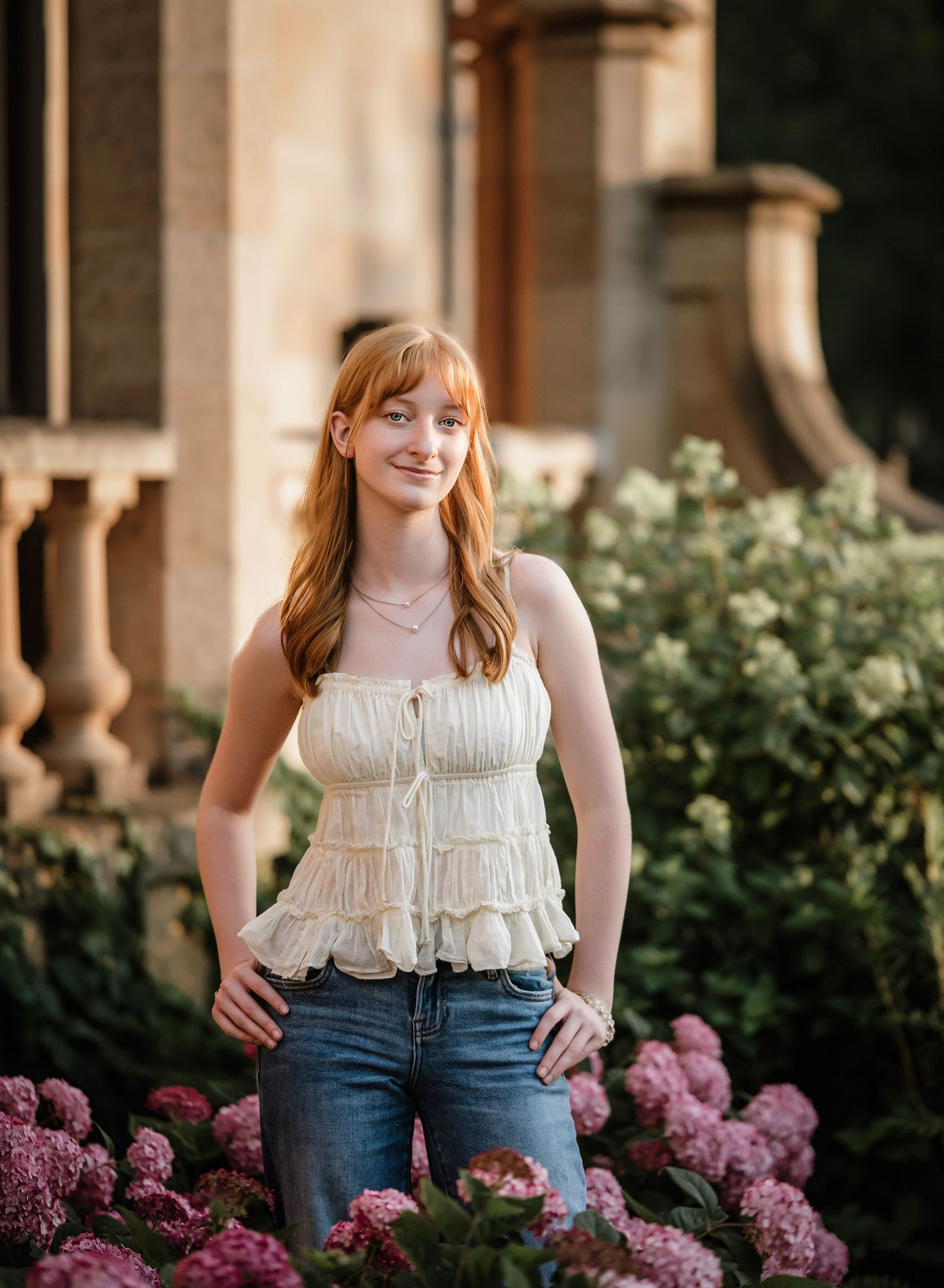 A person in a white ruffled top and jeans poses by purple hydrangea flowers in an outdoor garden setting.