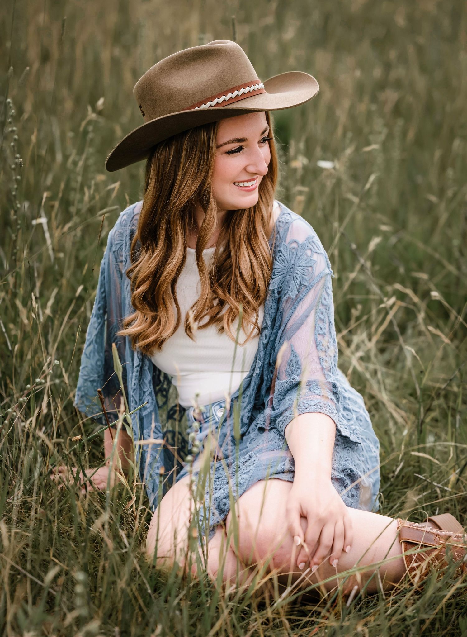 A person in a denim jacket and brown cowboy hat sits thoughtfully in a grassy meadow during golden hour.