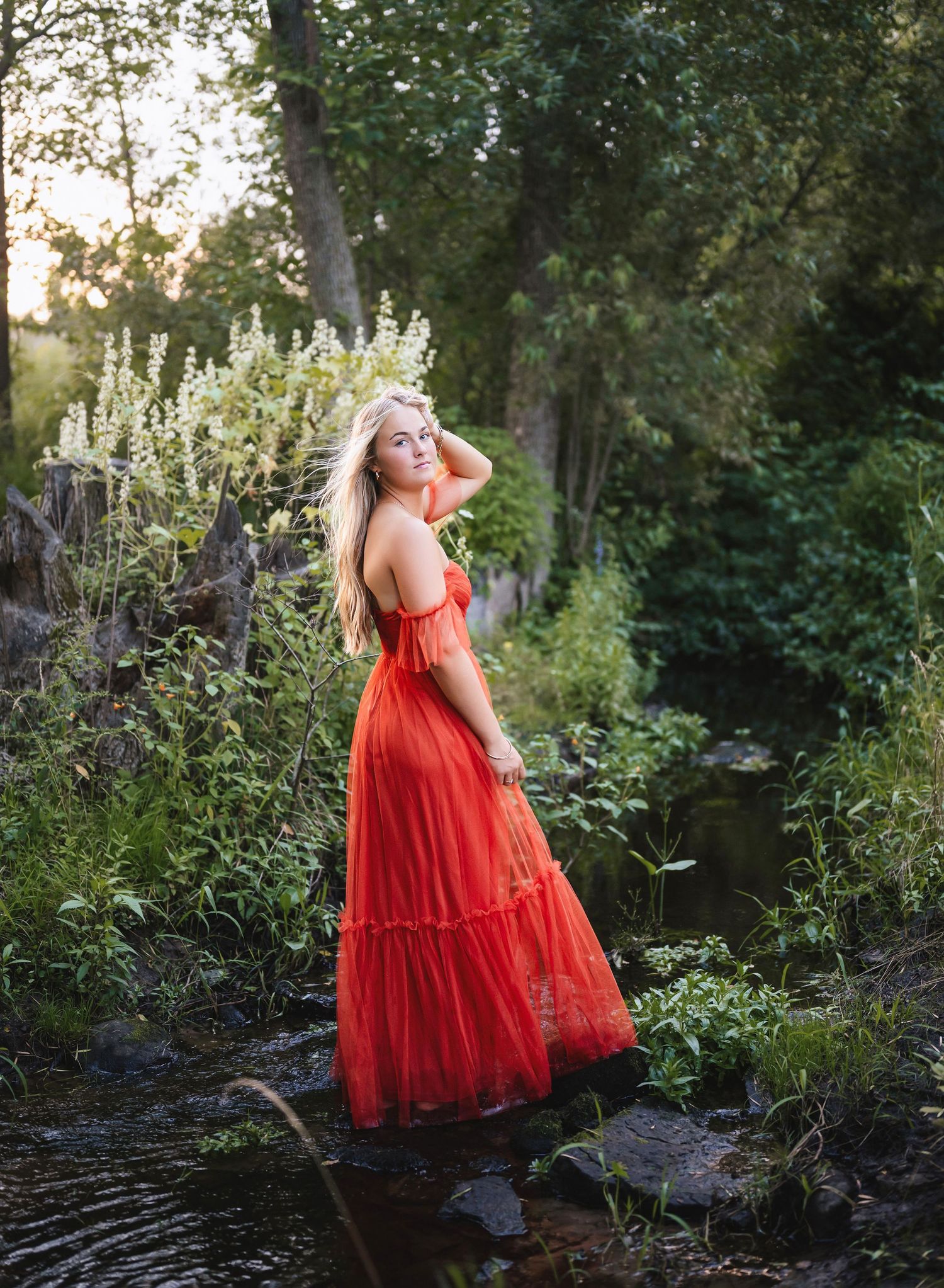 A figure in a flowing red dress poses gracefully by a stream in a forest during golden hour.