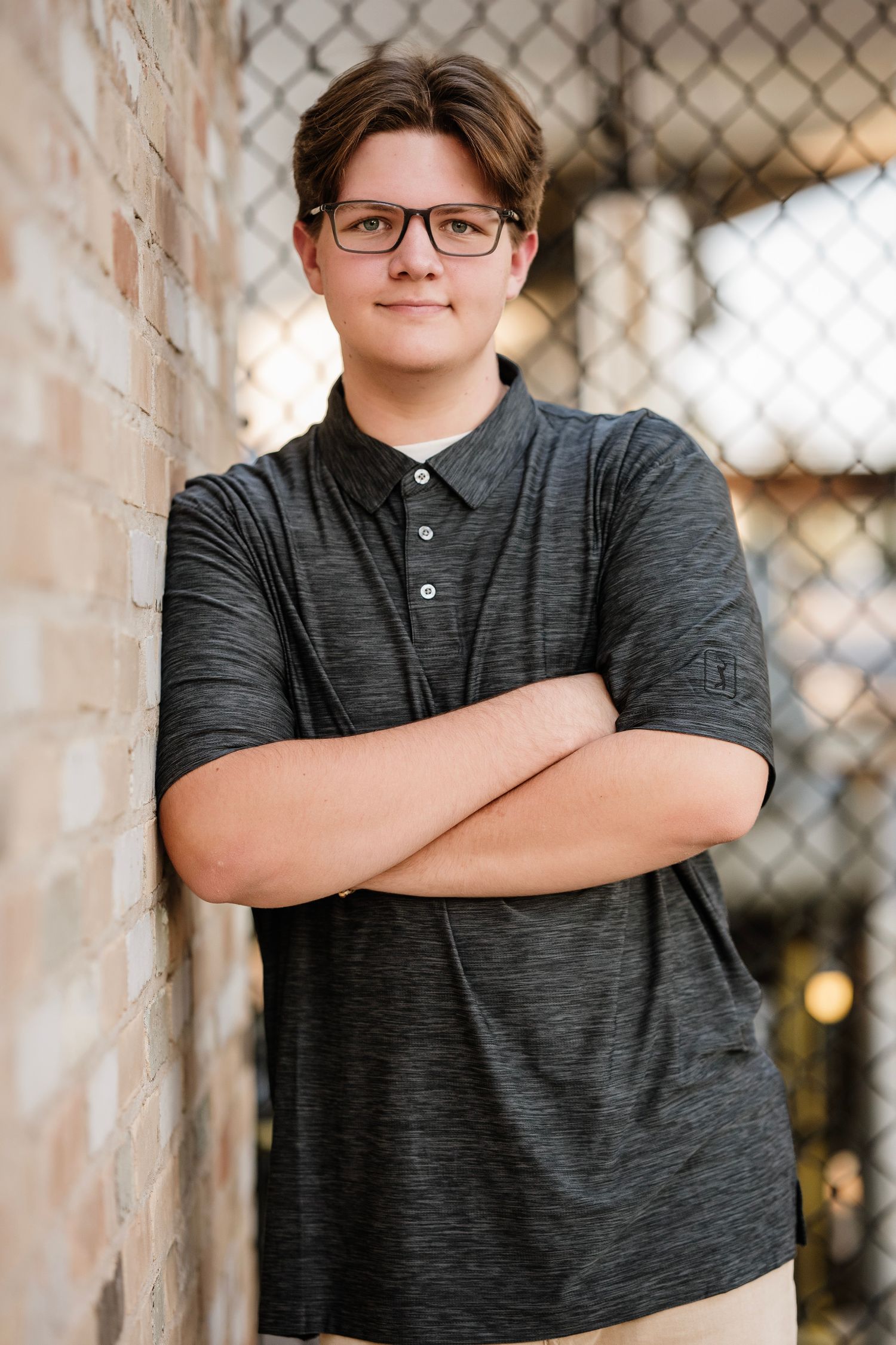 A student in glasses and a dark polo shirt stands with crossed arms against a brick wall with chain link fence in background.