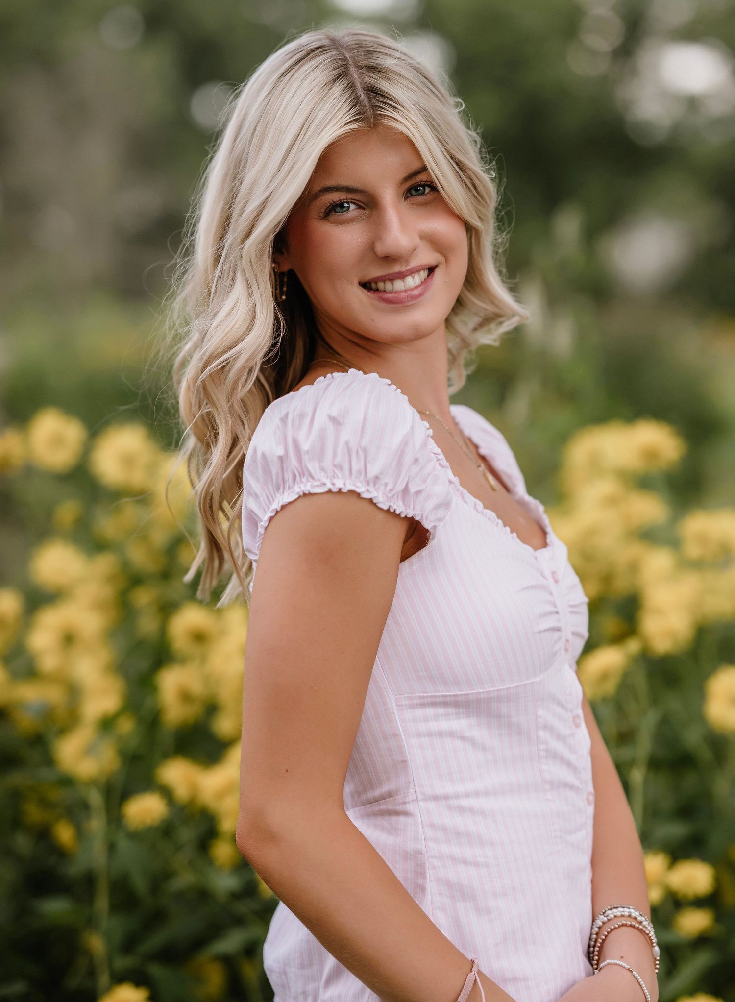 A person in a white ruffled blouse poses outdoors against a backdrop of yellow flowers in a garden setting.