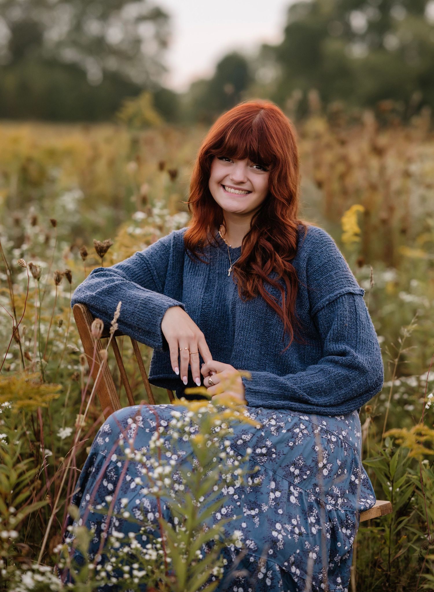 A person in a blue sweater and floral skirt sits gracefully among wildflowers in a sunlit meadow.