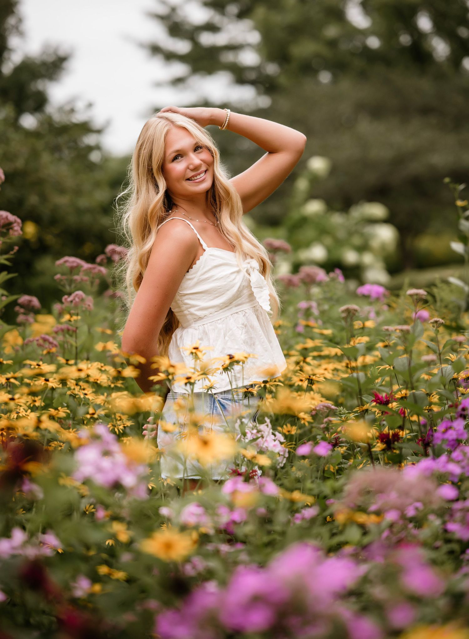 A person in a white dress poses gracefully among vibrant yellow and purple wildflowers in a summer garden setting.