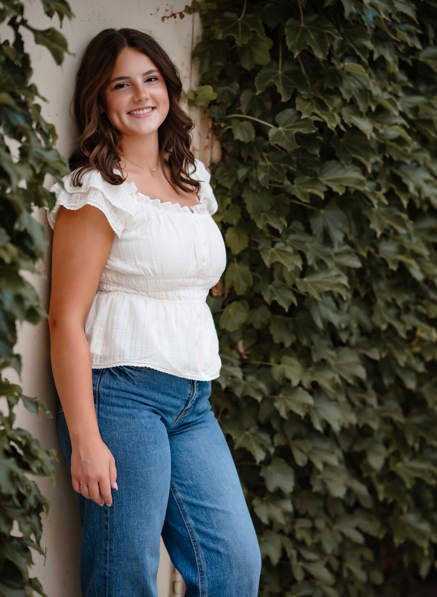A person wearing a white ruffled top and blue jeans poses against an ivy-covered wall in an outdoor portrait setting.