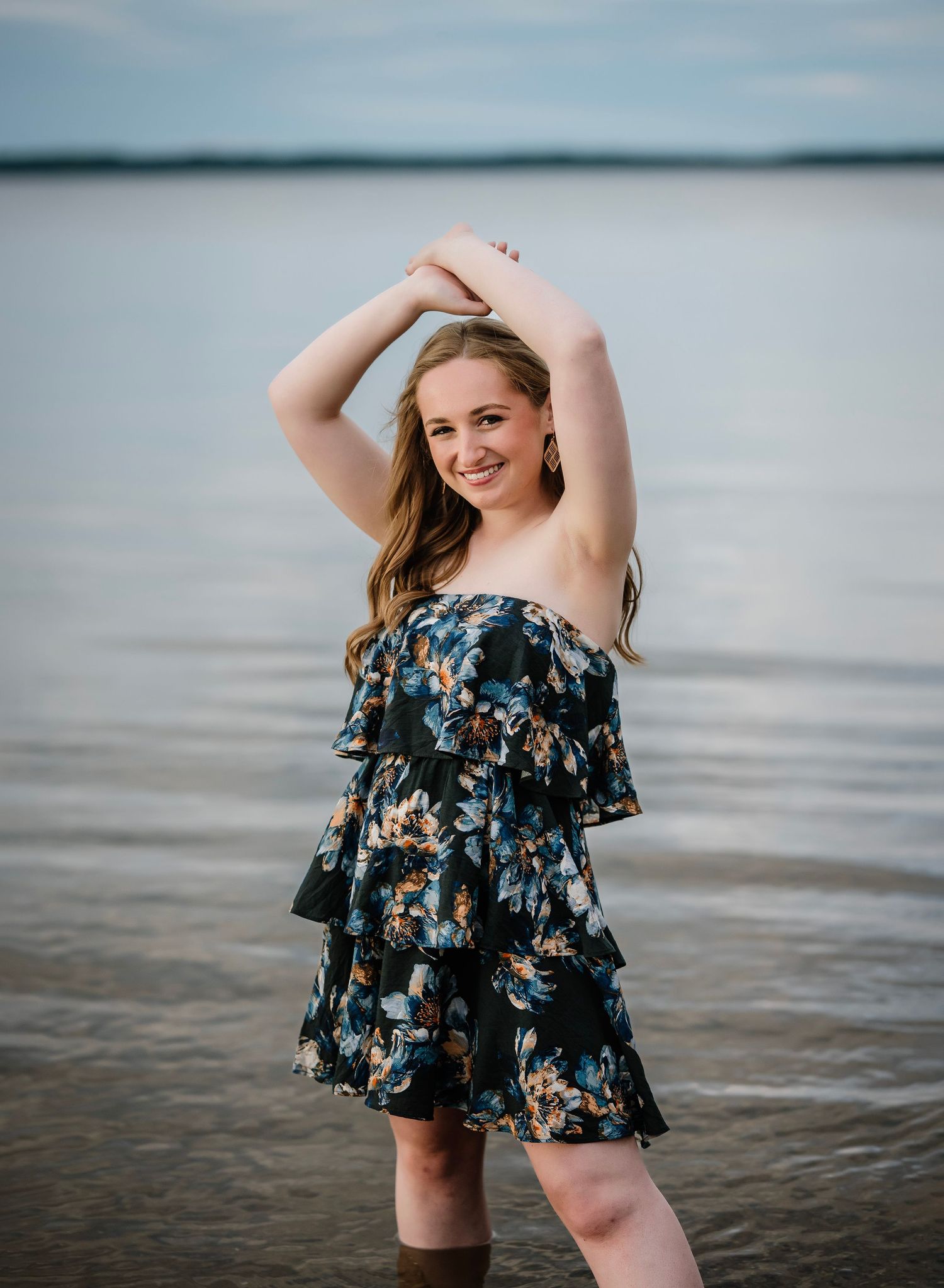 Young woman in floral off-shoulder dress posing gracefully at a lake shore with hands raised above her head.